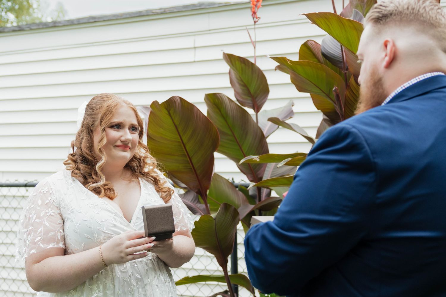 Bride smiles at groom while presenting a ring box during an outdoor first look.