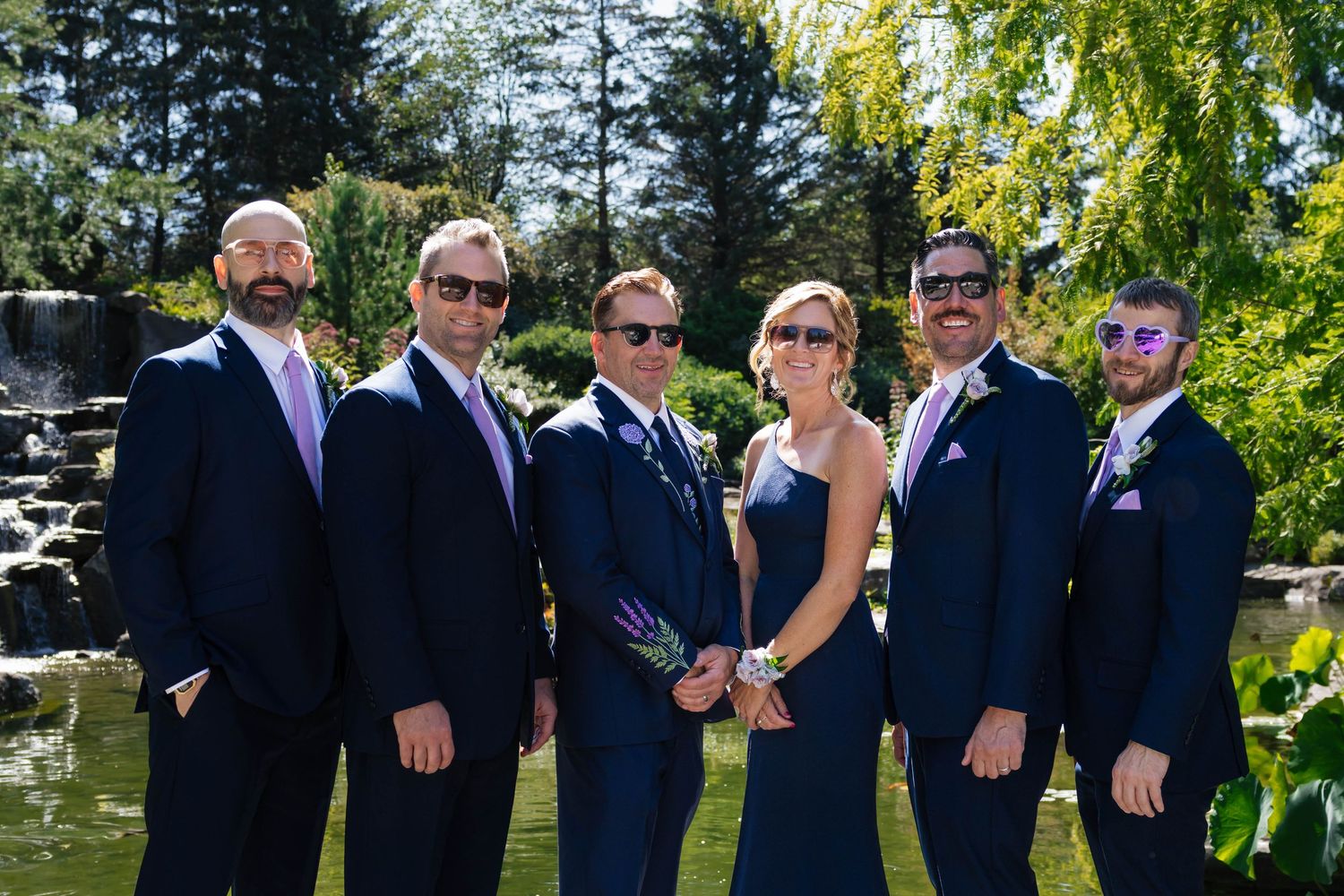 Groomsmen and groomswoman in navy suits with lavender boutonnieres pose together by garden pond on wedding day.