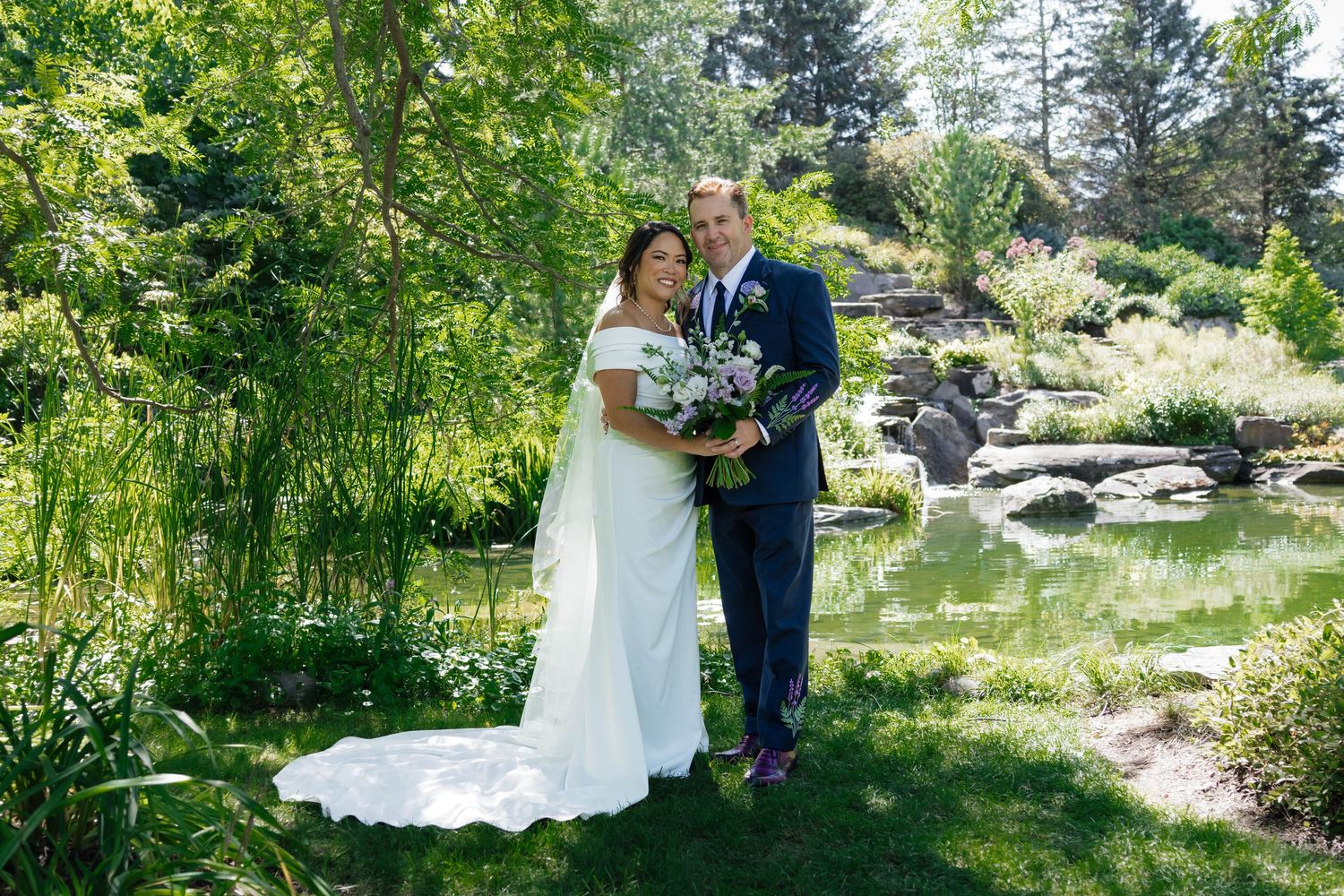 Bride and groom posing together near a serene garden pond surrounded by lush greenery.