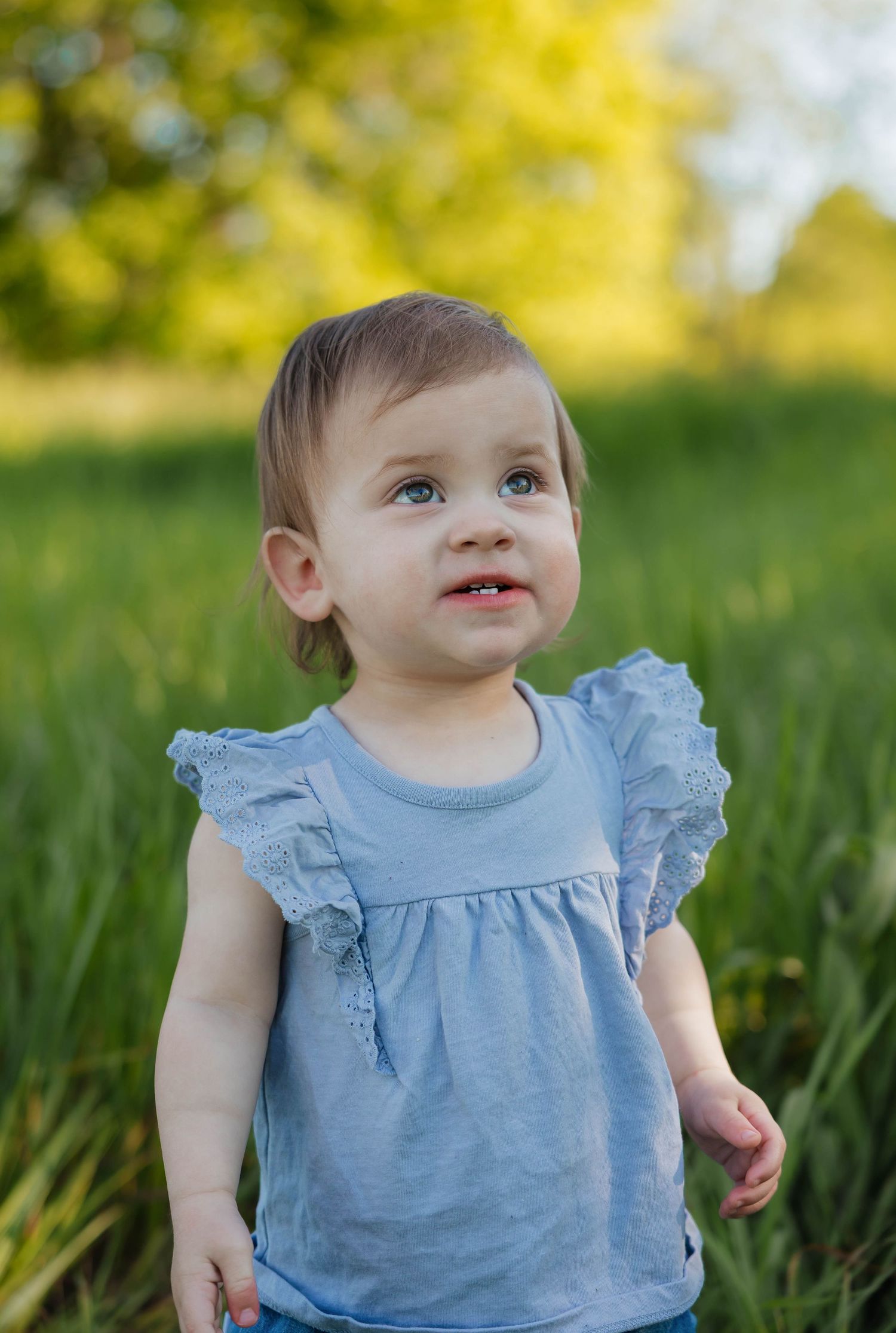 Sweet baby girl in a blue ruffle top standing in a green grassy field on a sunny day.