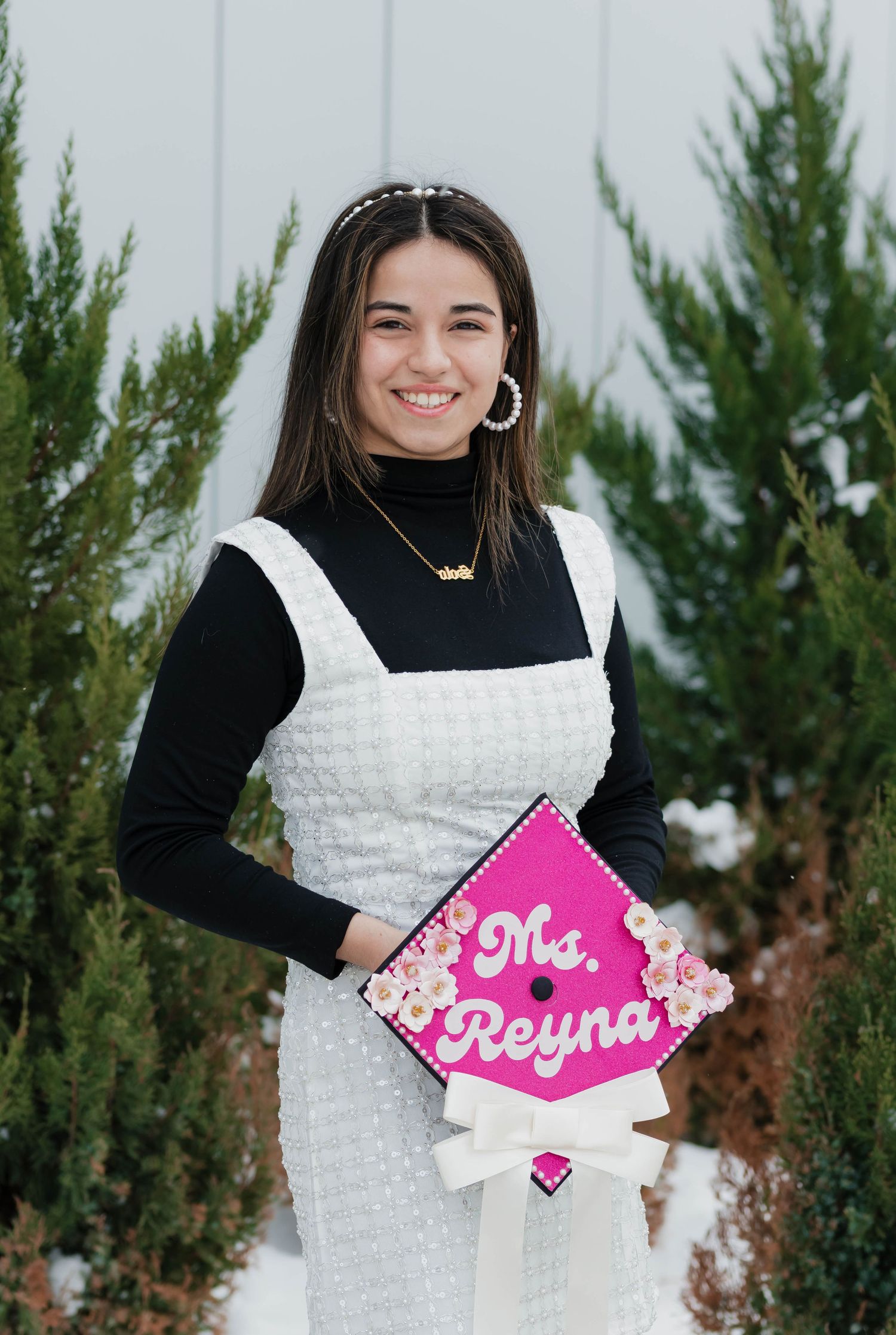 Smiling graduate holding pink Ms. Reyna decorated cap wearing white dress near evergreen trees.