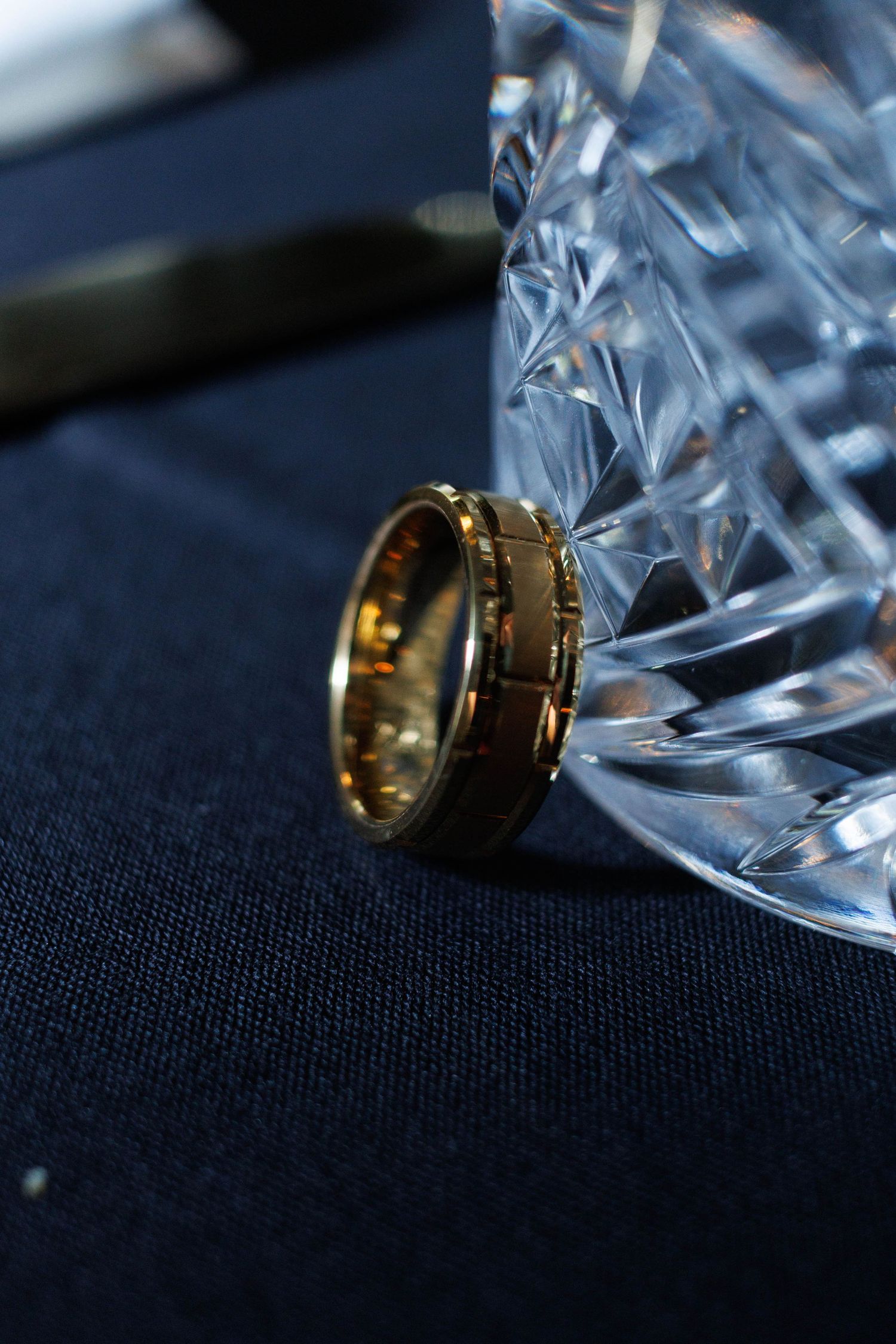 Gold wedding band ring leaning against a crystal glass on a dark navy blue tablecloth.