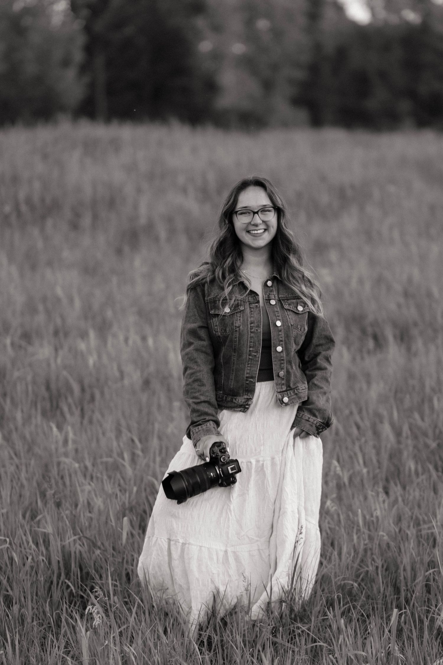 Black and white photo of a smiling woman holding a camera in a field, wearing a denim jacket and white skirt.