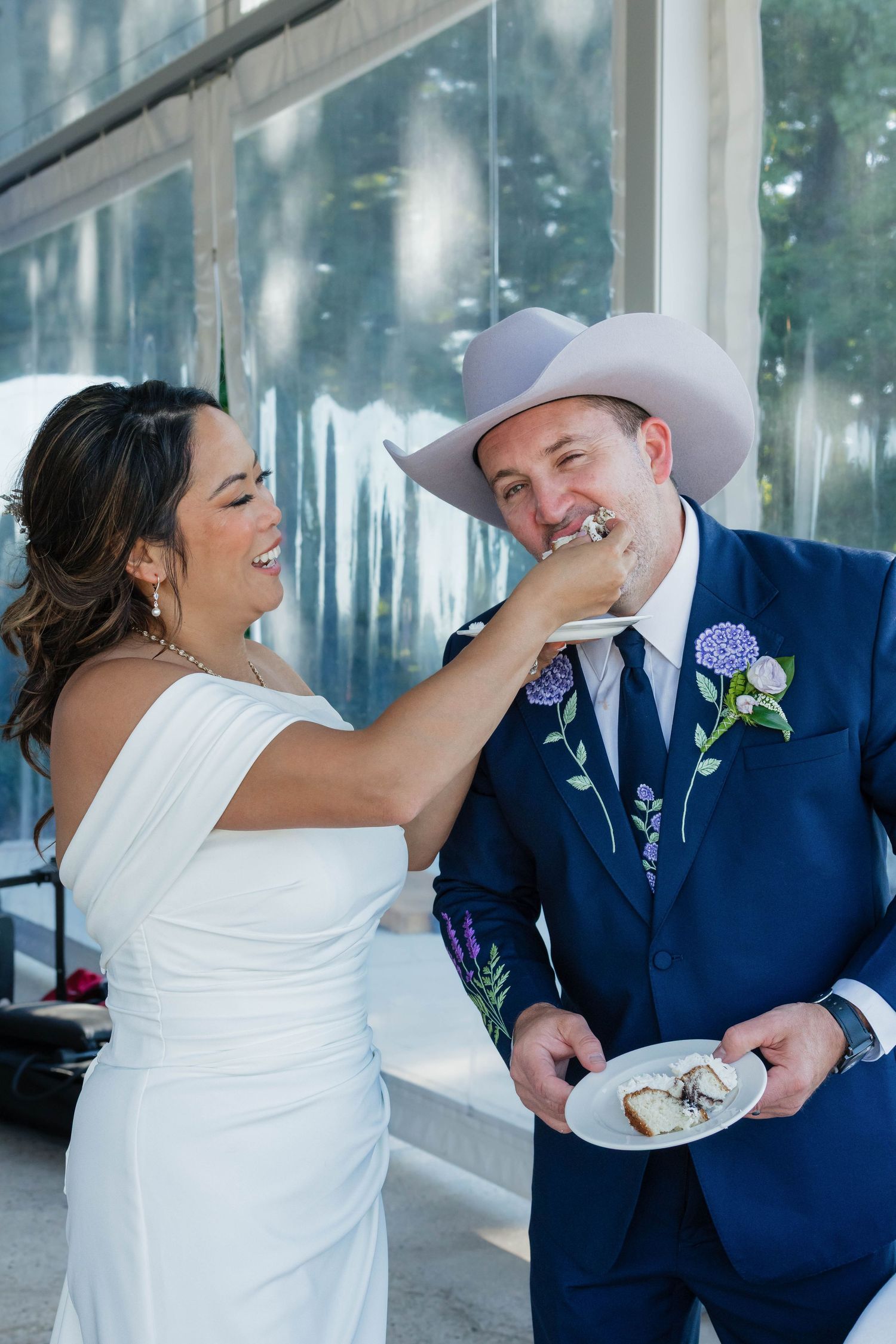 Smiling bride feeds wedding cake to groom wearing navy suit and cowboy hat at reception.