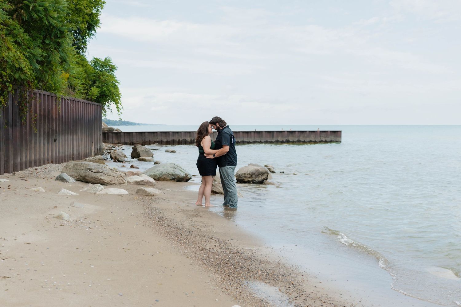A couple shares a romantic kiss on a sandy beach near calm water and a wooden pier.