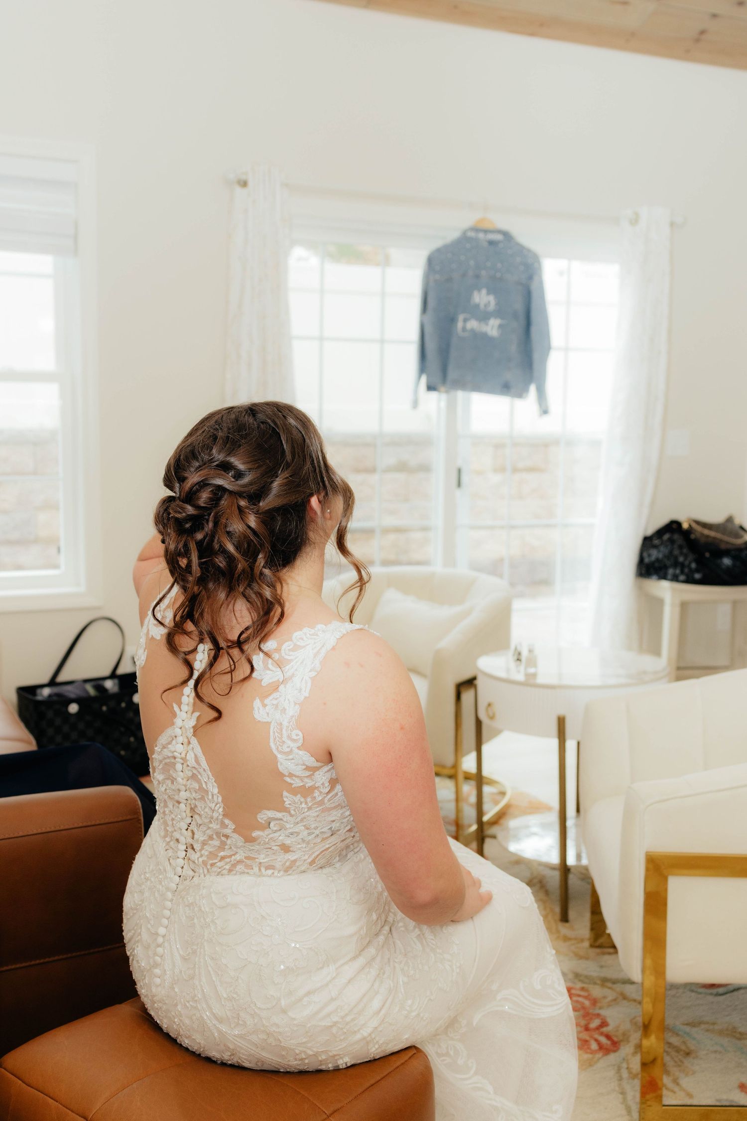 Bride in lace wedding gown sits facing window with personalized denim jacket hanging nearby.