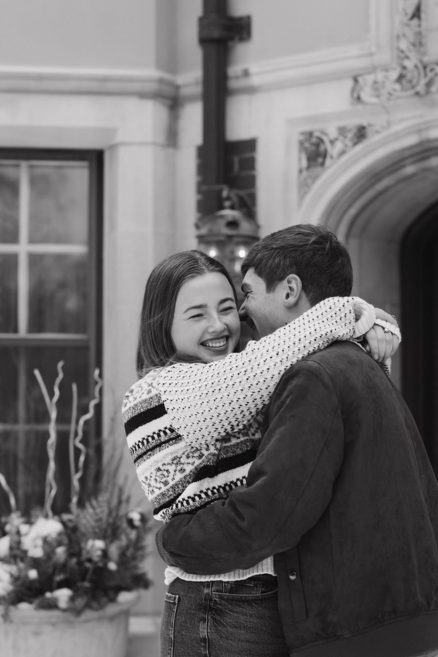 Black and white photo of a joyful couple hugging and laughing outside a grand building.