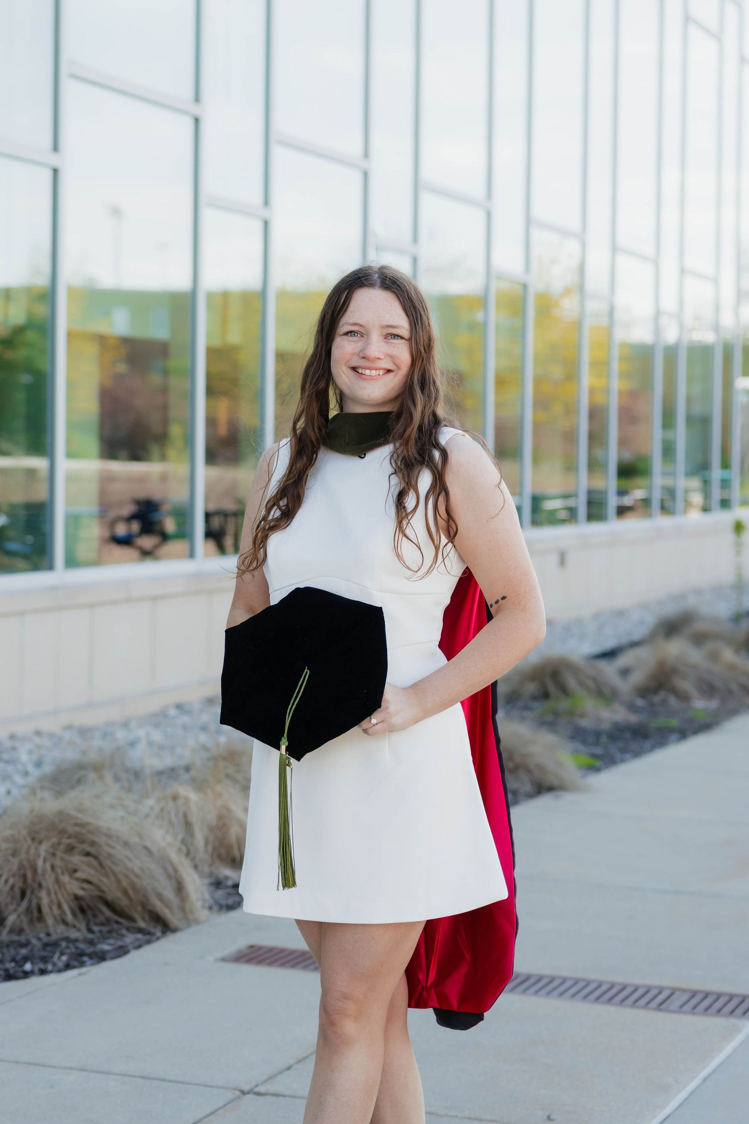 Smiling graduate in white dress holds black mortarboard cap with gold tassel outside building.