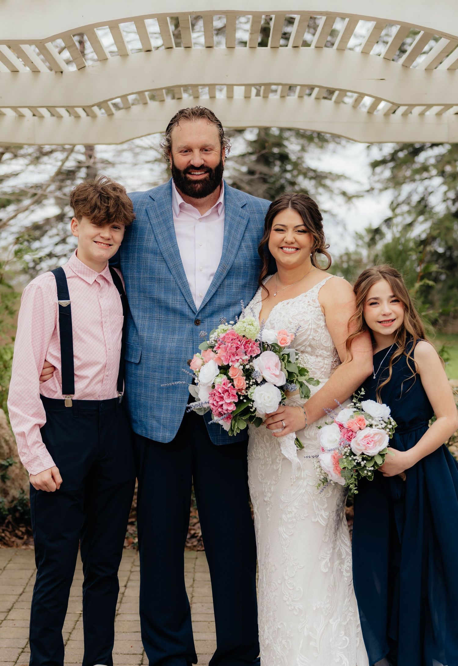 Bride with pink bouquet poses with groom in blue plaid suit and two children under garden arbor.