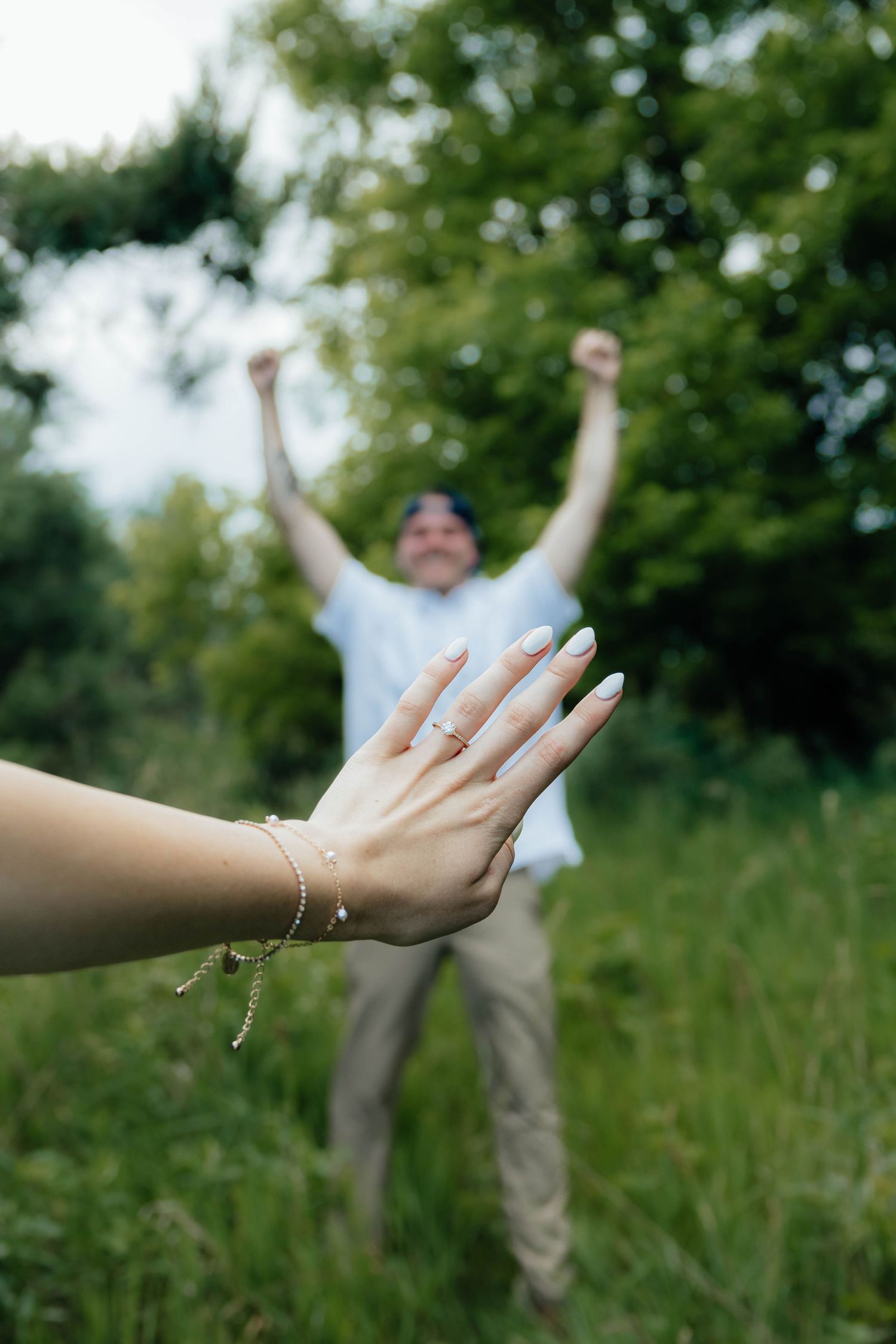 Woman displaying engagement ring in foreground while excited man celebrates with raised arms behind her.