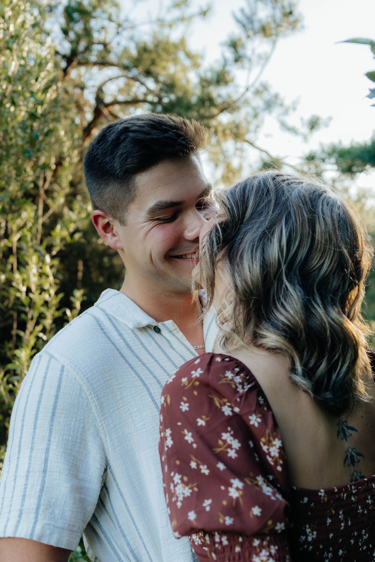 Couple sharing an intimate moment outdoors, man smiling as woman leans in close against a natural green backdrop.