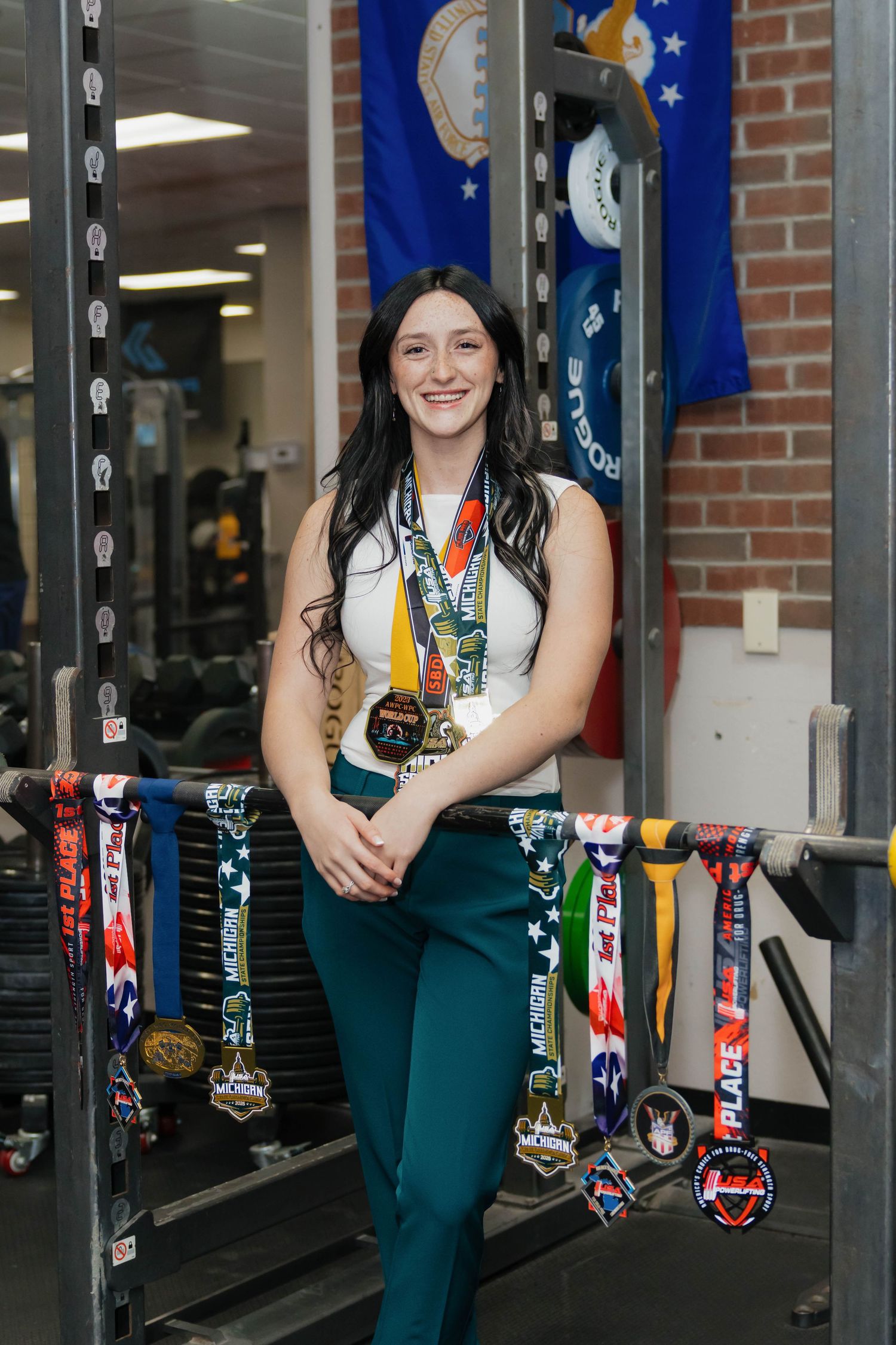 Young woman posing with multiple sports medals in a gym with military flags in the background.