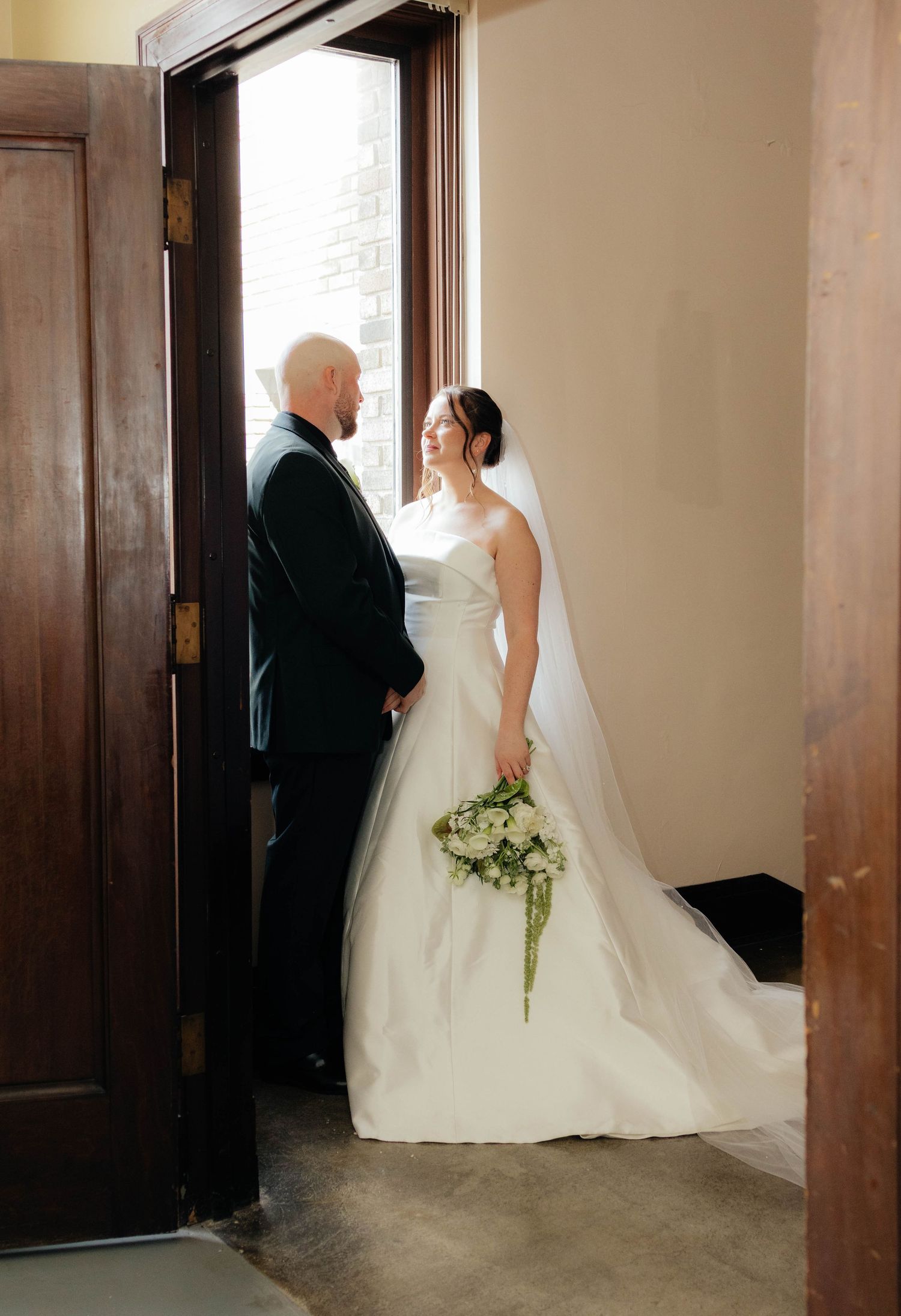 Bride and groom share an intimate moment by a doorway, bride holds white bouquet.