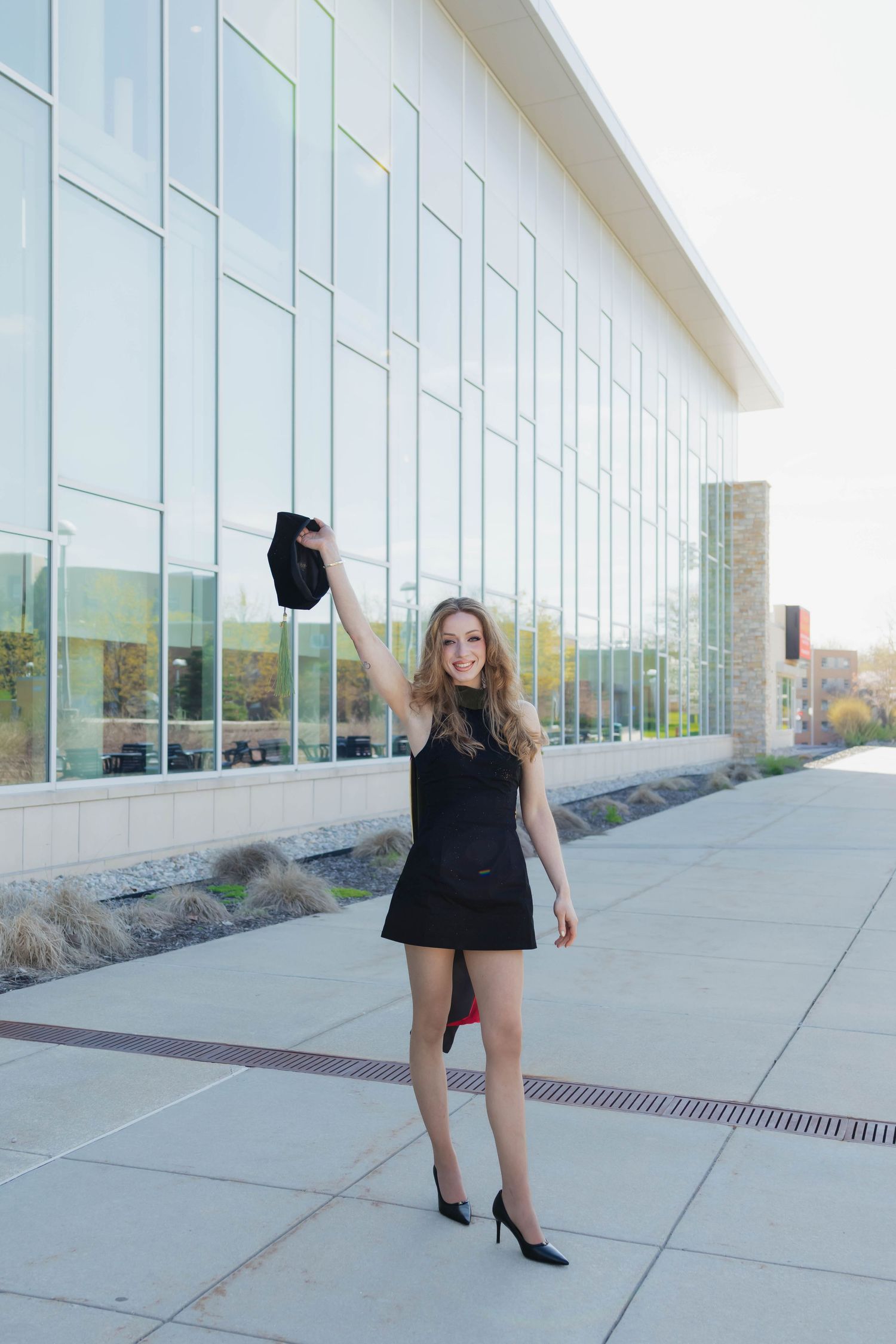 Young woman in black mini dress raising graduation cap triumphantly outside modern university building.