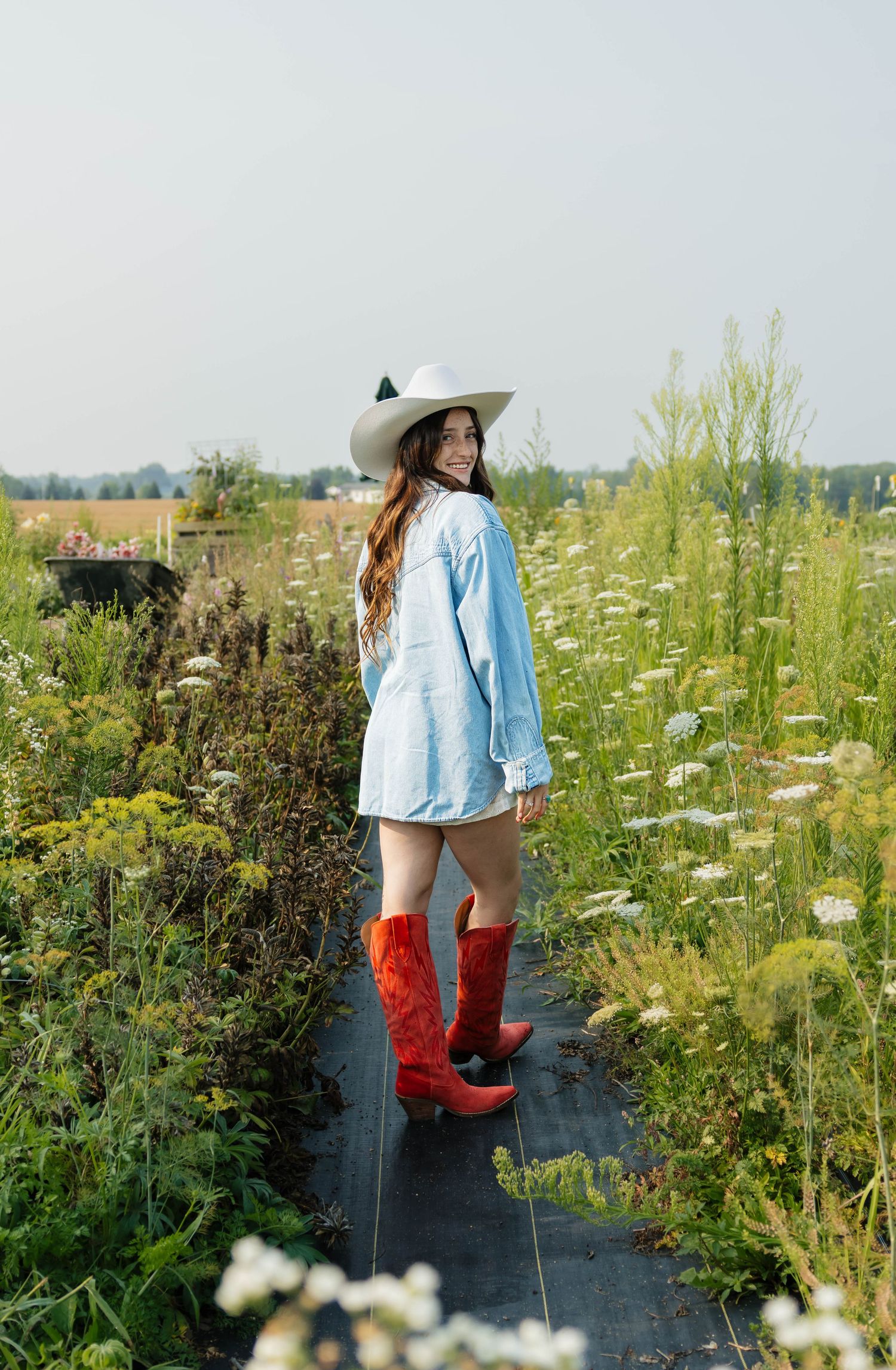 Teen girl in denim jacket, white hat, and red cowboy boots posing in lush flower field.