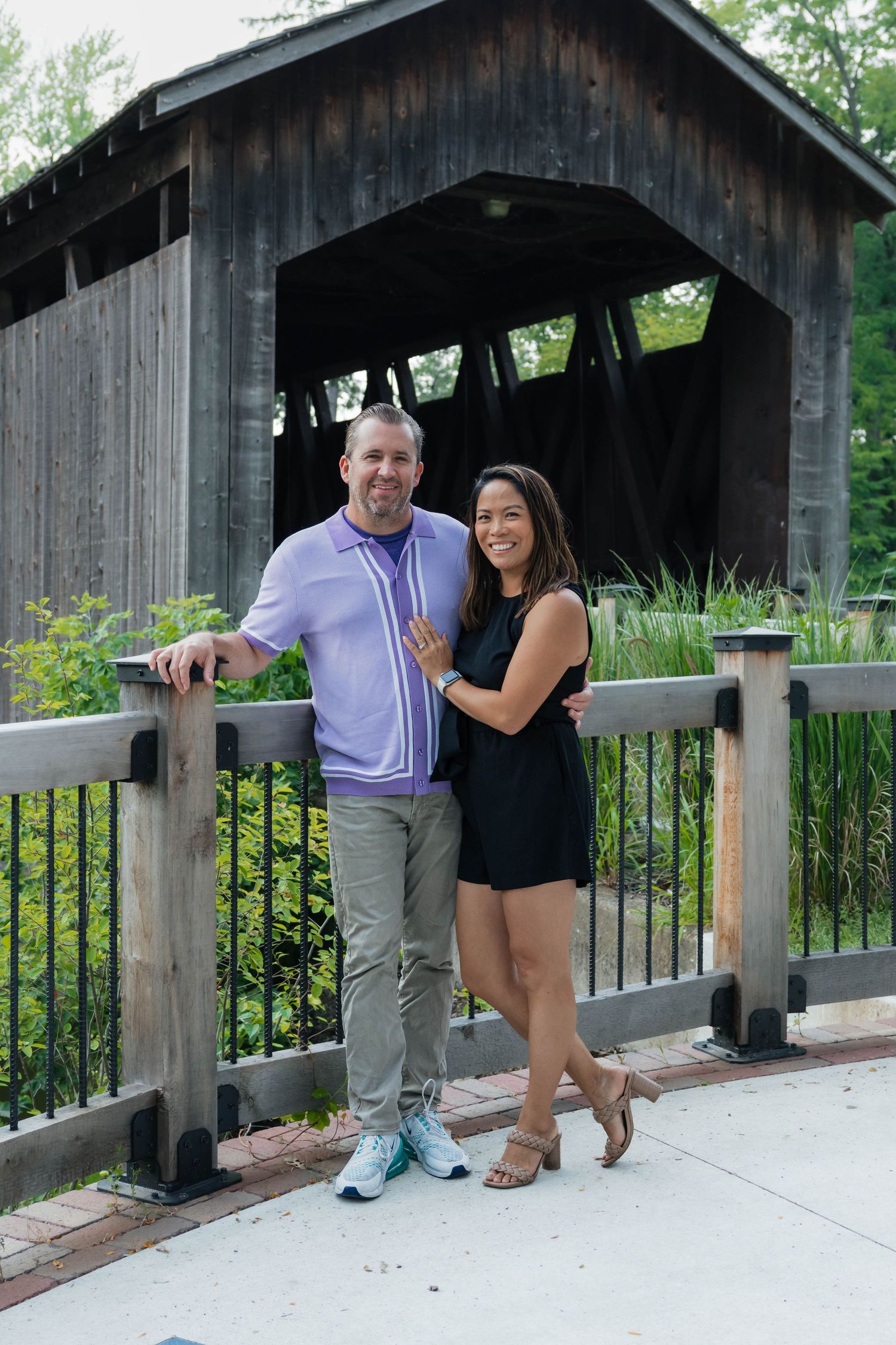 Couple smiling together in front of a rustic covered bridge outdoors.