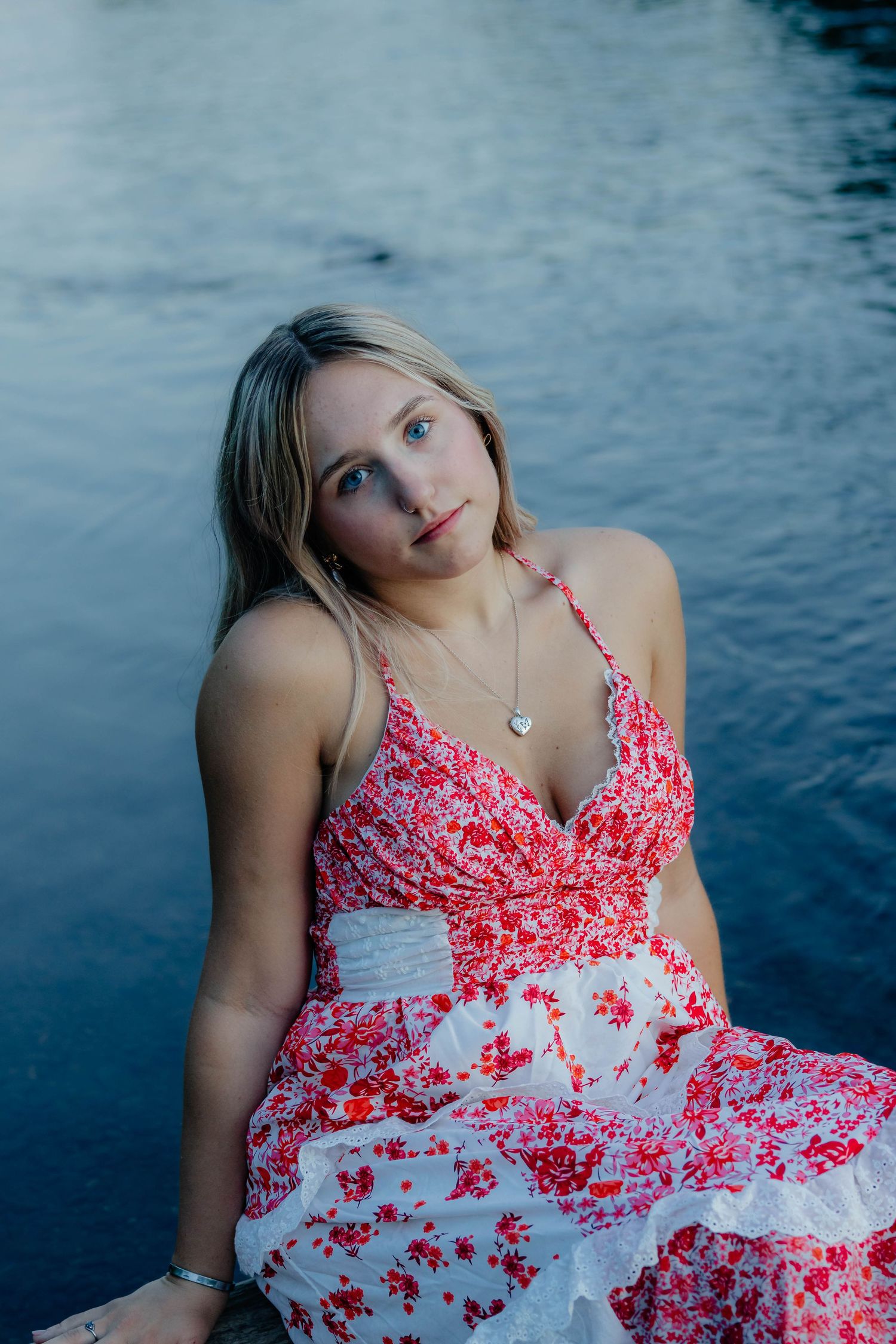 Young blonde woman in red floral dress sitting by water.