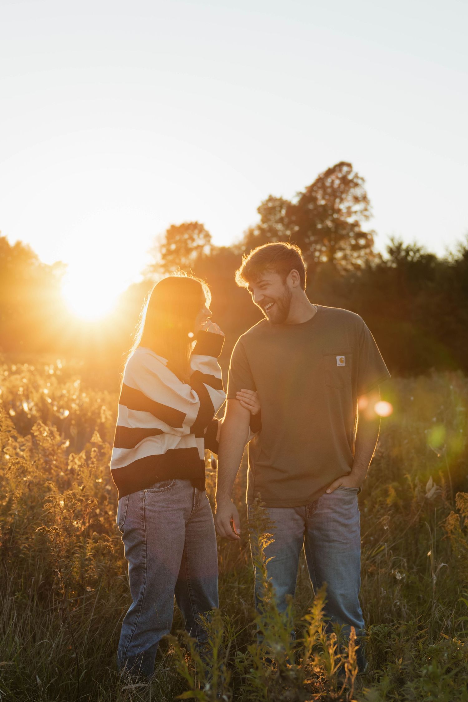 Couple laughing together in a glowing sunlit meadow during a beautiful golden hour sunset.