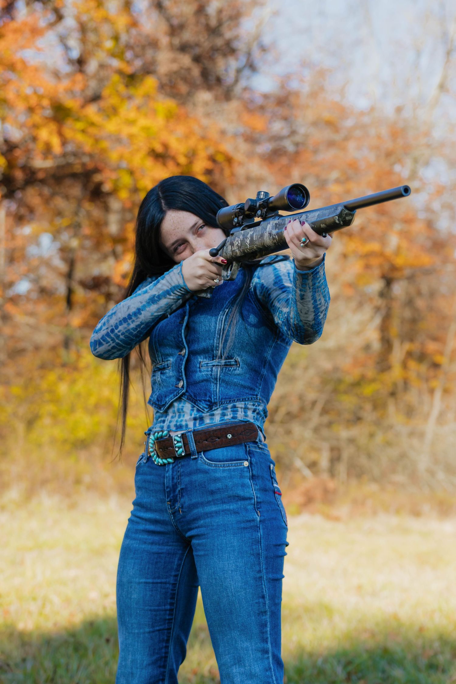 Woman in denim outfit aiming a camouflage rifle with scope in an autumn forest setting.