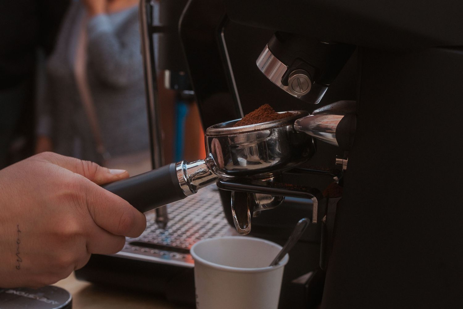 A barista uses an espresso machine portafilter handle with a coffee cup and paperwork on a dark wooden counter.