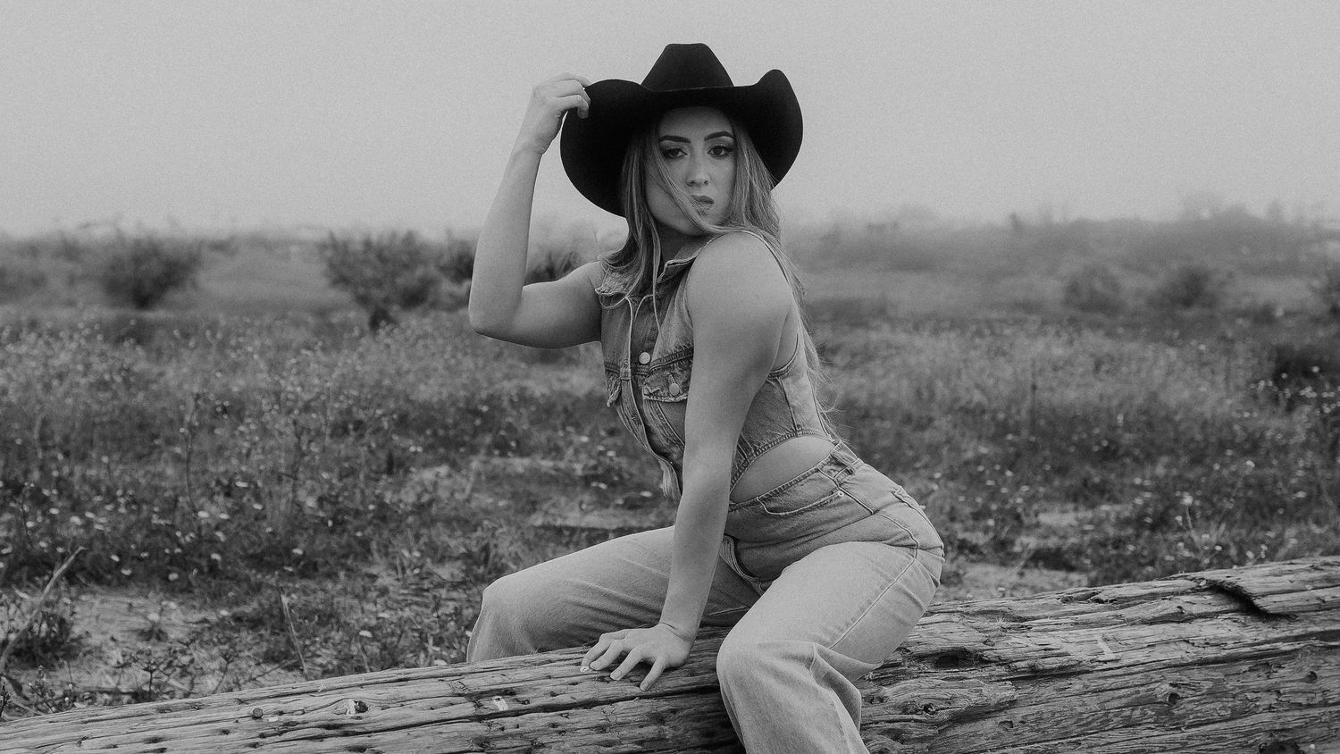Black and white portrait of a person in a cowboy hat posing on a rustic log in a rural setting.