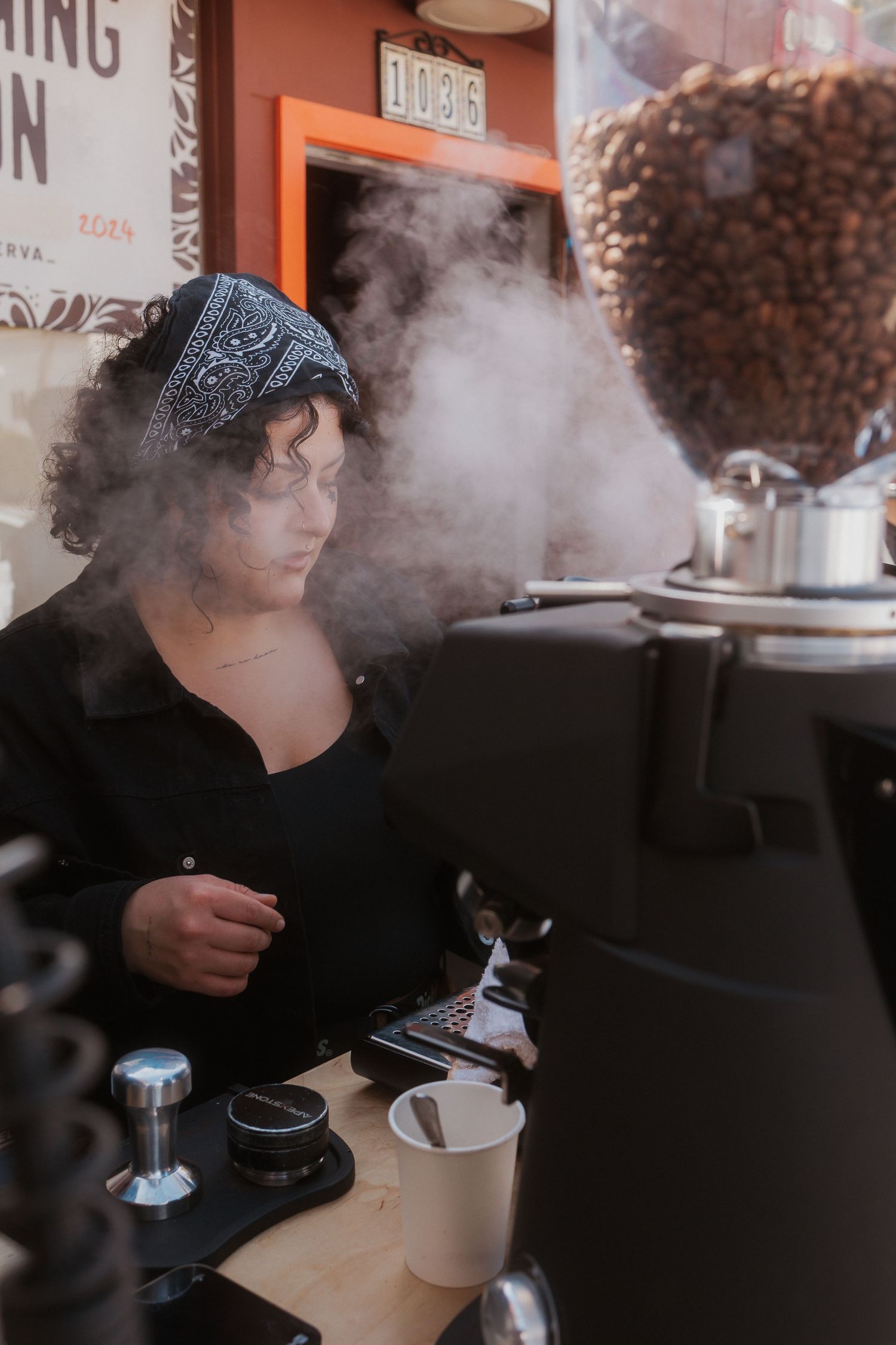 Barista preparing an espresso with steam rising from coffee equipment and cups in a moody cafe setting.