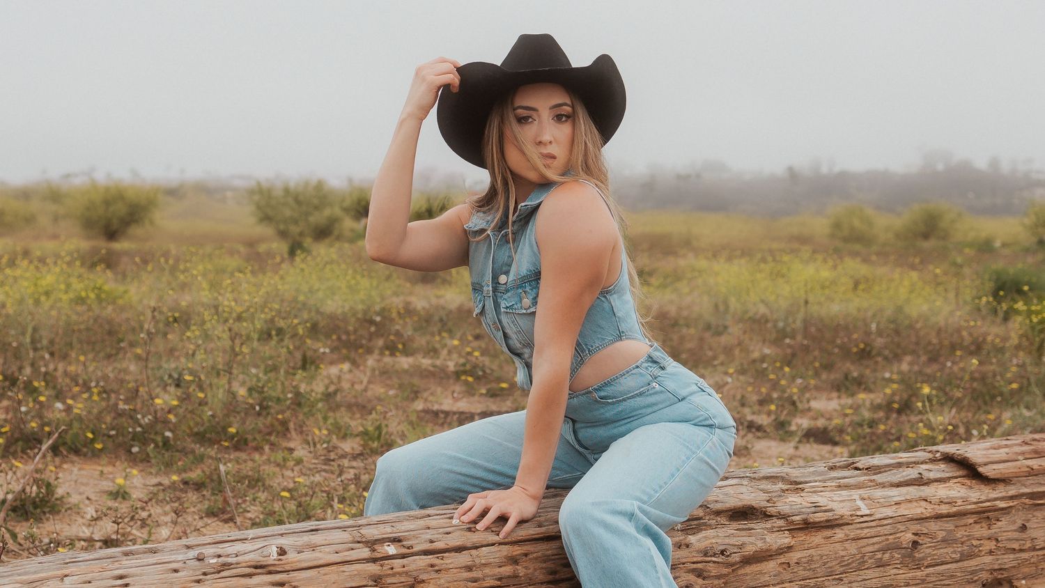 A woman in a cowboy hat and denim overalls poses on a fallen log in a rustic outdoor setting at sunset.