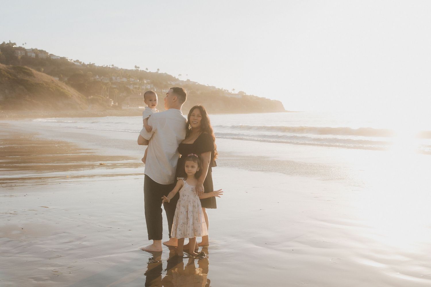 Family of four walking along a beach at sunset with sunlit waves and sand creating a dreamy coastal atmosphere.