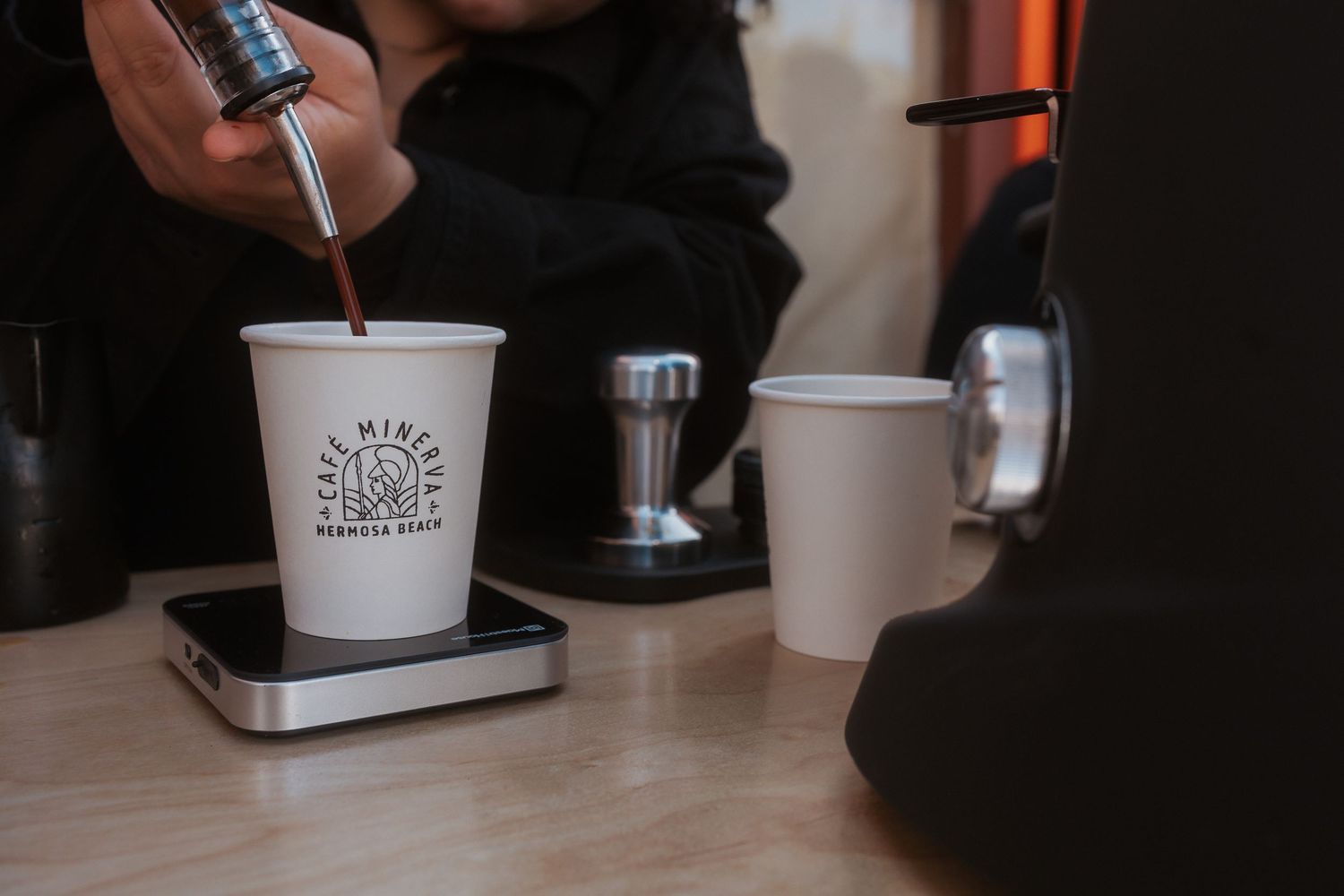 Barista pouring a drink into a branded white paper cup on a wooden surface next to additional coffee cups.