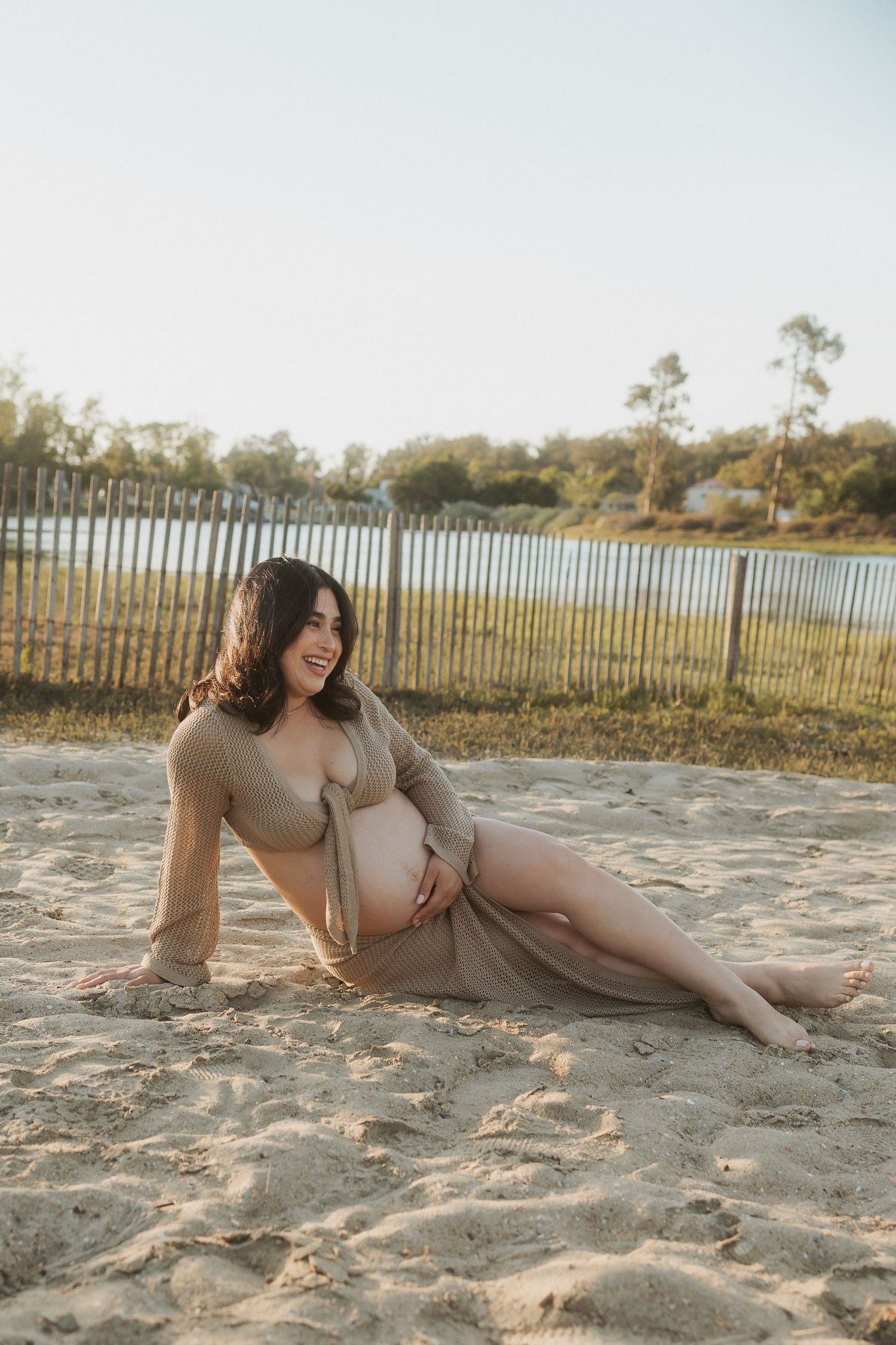 Expectant woman in tan dress relaxes on sandy beach near lake during golden hour with fence in background.