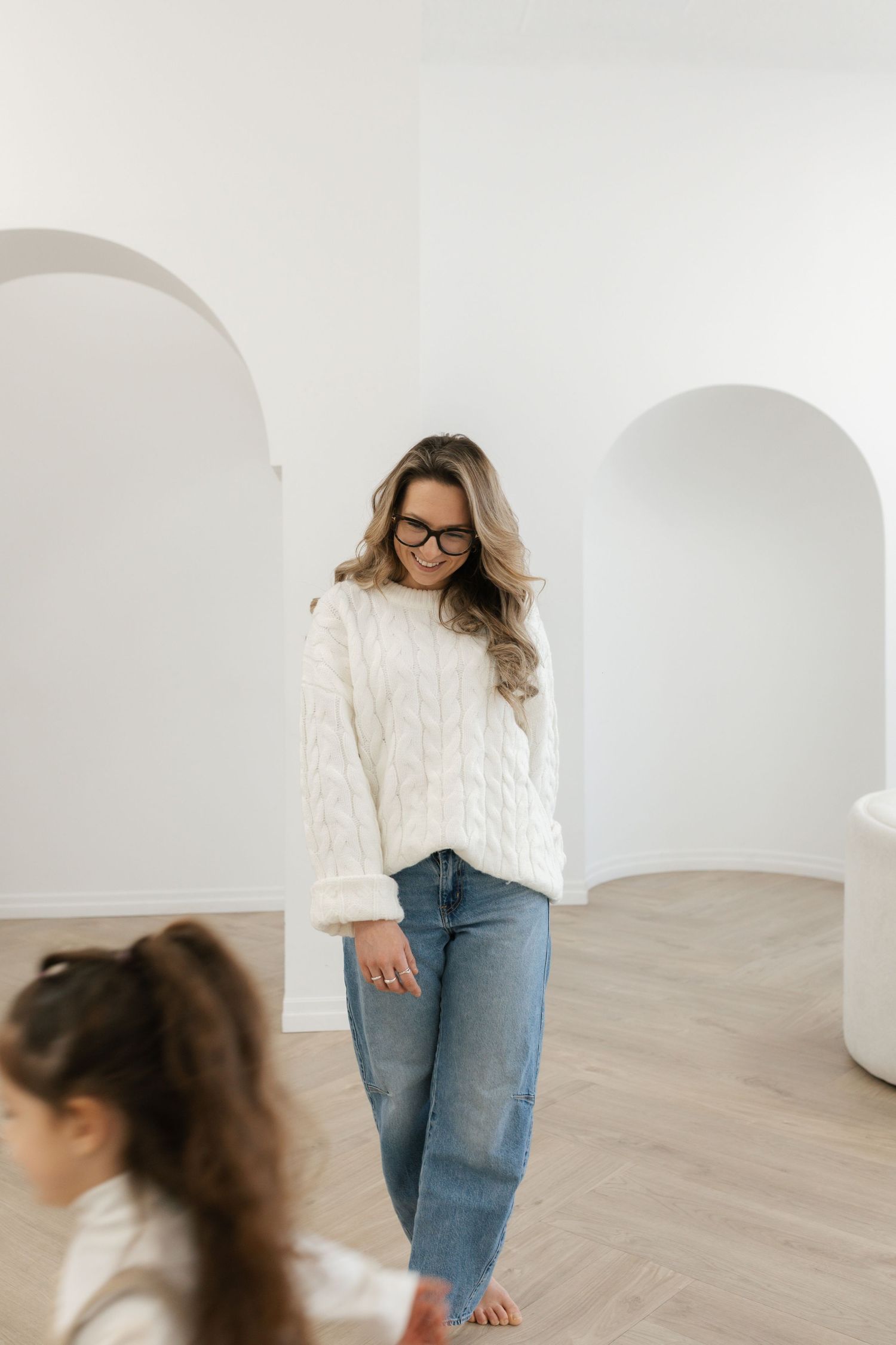 Woman in white cable-knit sweater and jeans wearing glasses stands in bright minimalist room with arched doorways.