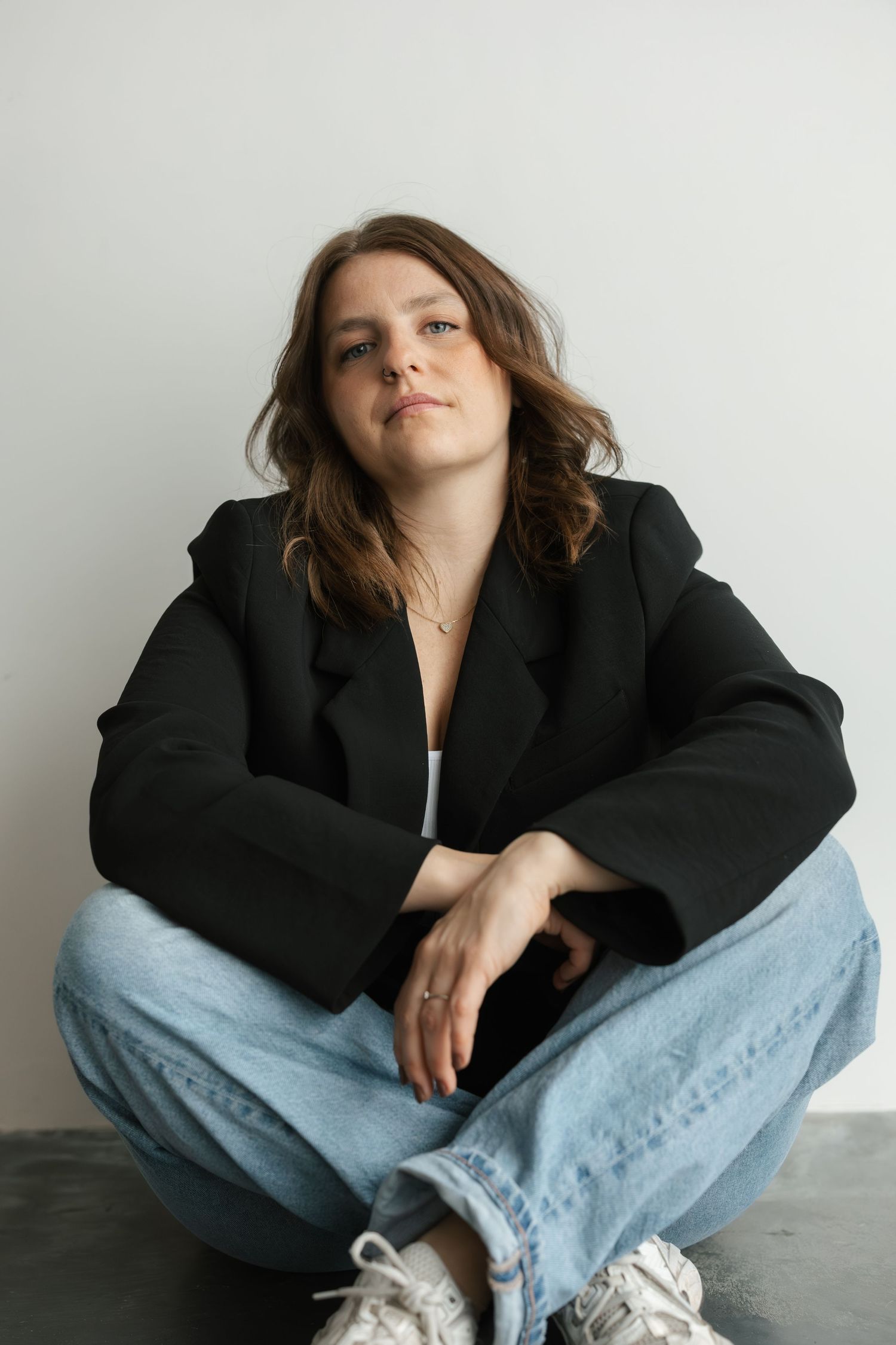 A young woman in a black blazer and jeans sits cross-legged on the floor against a white wall looking at camera.