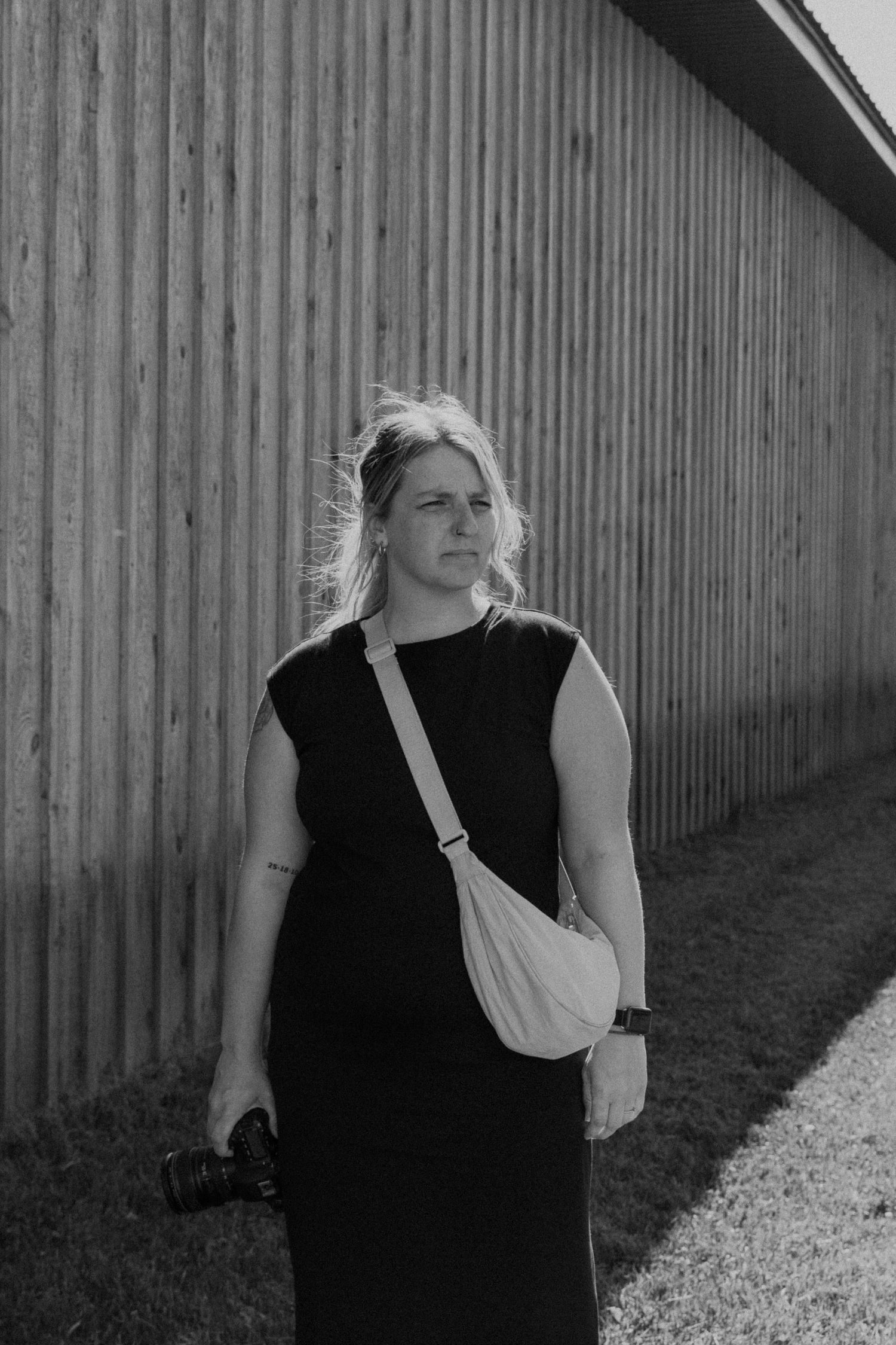 Black and white portrait of a female photographer in black dress carrying camera along a wooden fence.