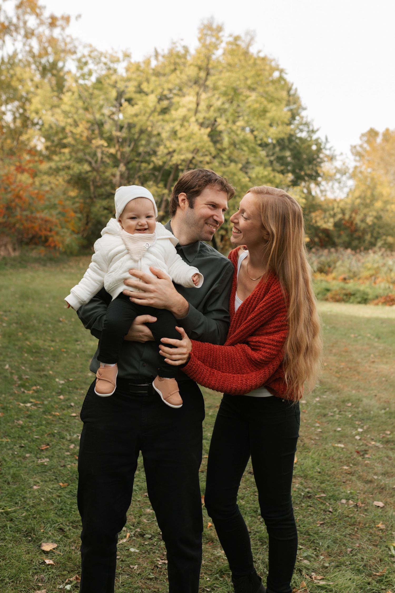 A happy young family with a smiling baby in white jacket poses outdoors in a colorful autumn park setting.