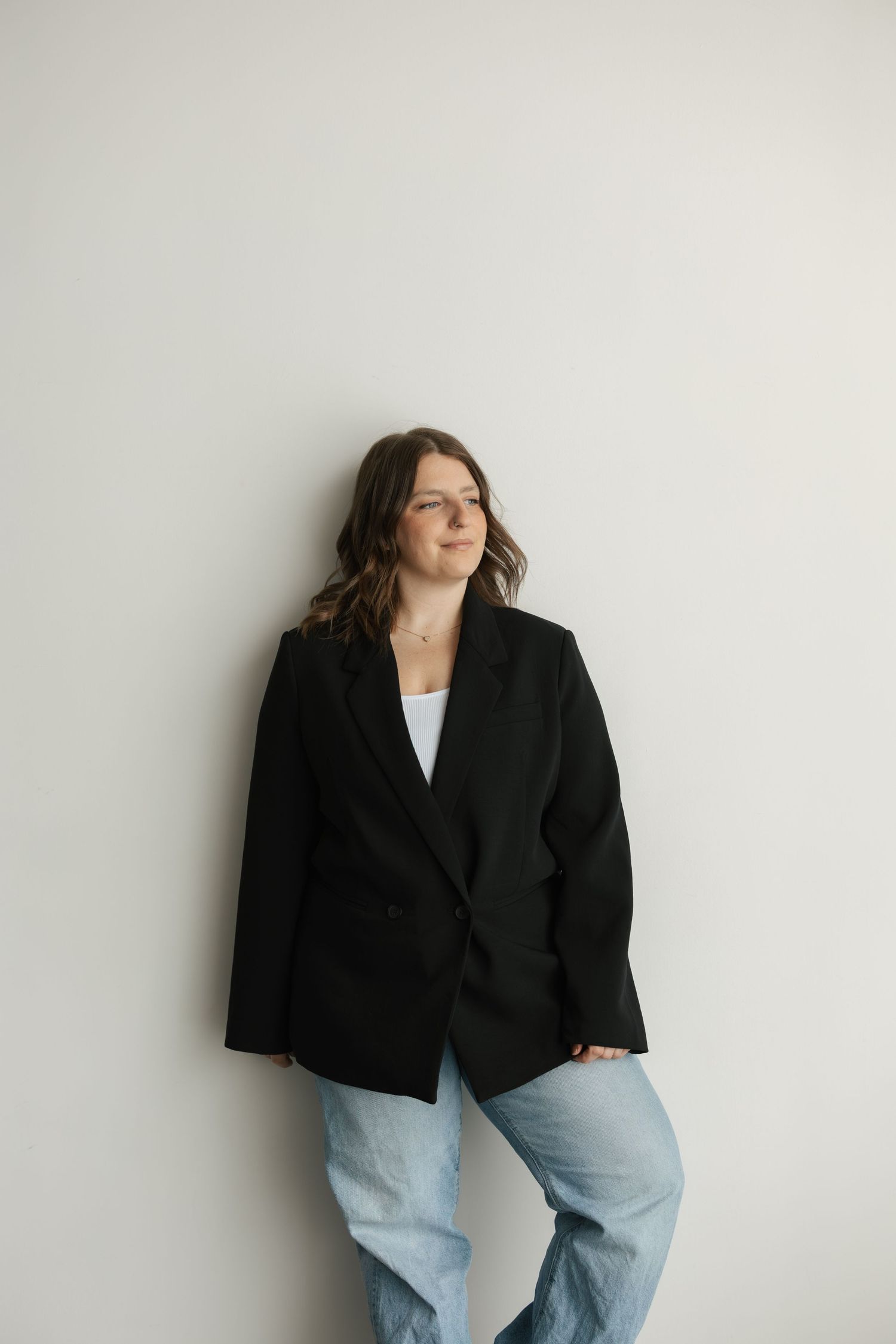 A young woman in a black oversized blazer and light blue jeans leans against a white wall looking to the side.