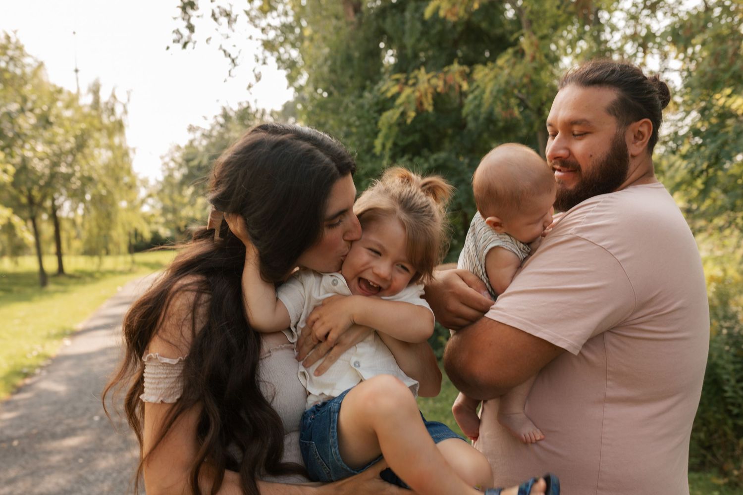 Happy family of four enjoying a sunny day outdoors, parents kissing and holding their toddler and baby in a green park.