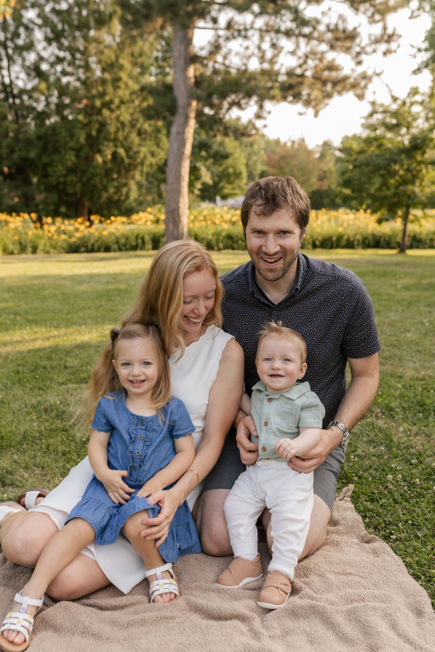 Happy family of four sitting on a blanket in a sunny park, parents smiling with young daughter and baby son.