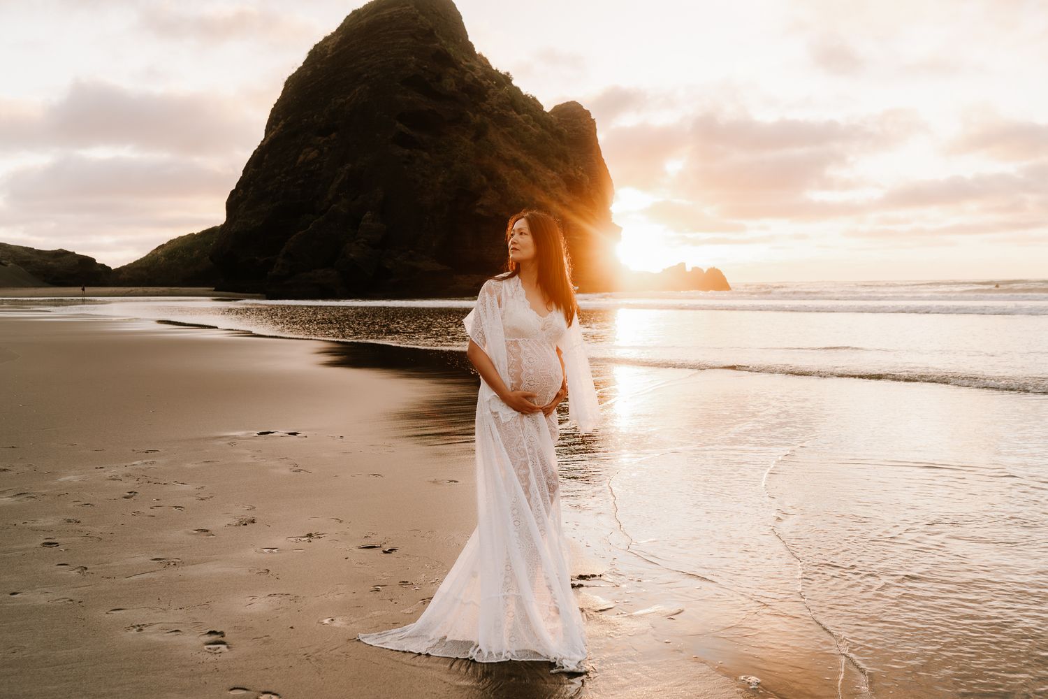 woman is wearing a white dress and is holding her baby belly as the sun sets behind her on the beach