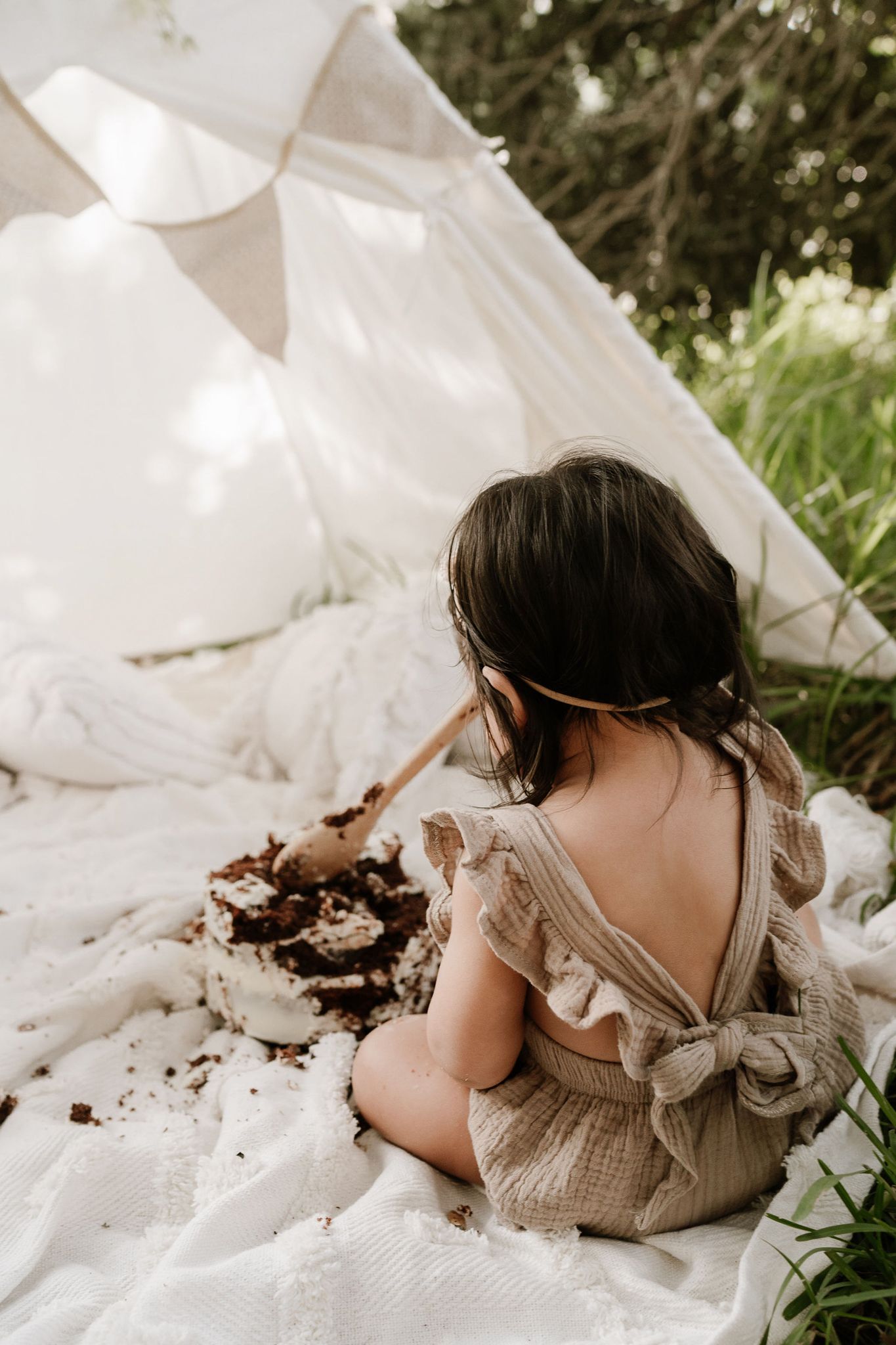 Outdoor cake smash session with a baby seated in front of a teepee, surrounded by long grass. A whimsical, natural setting with playful, mes