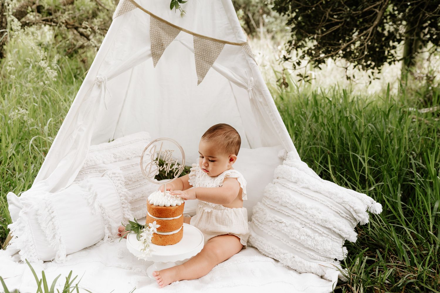 Outdoor cake smash session with a baby seated in front of a teepee, surrounded by long grass. A whimsical, natural setting with playful, mes