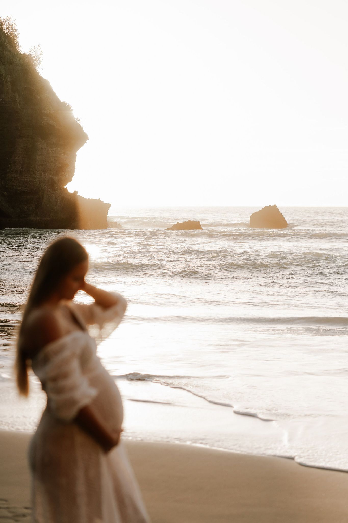woman is wearing a white dress and is holding her baby belly as the sun sets behind her on the beach