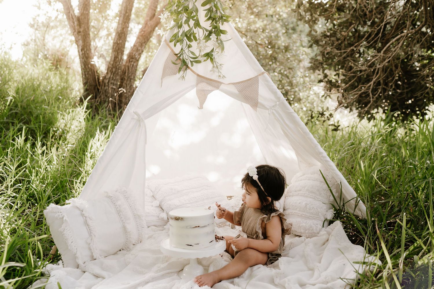Outdoor cake smash session with a baby seated in front of a teepee, surrounded by long grass. A whimsical, natural setting with playful, mes