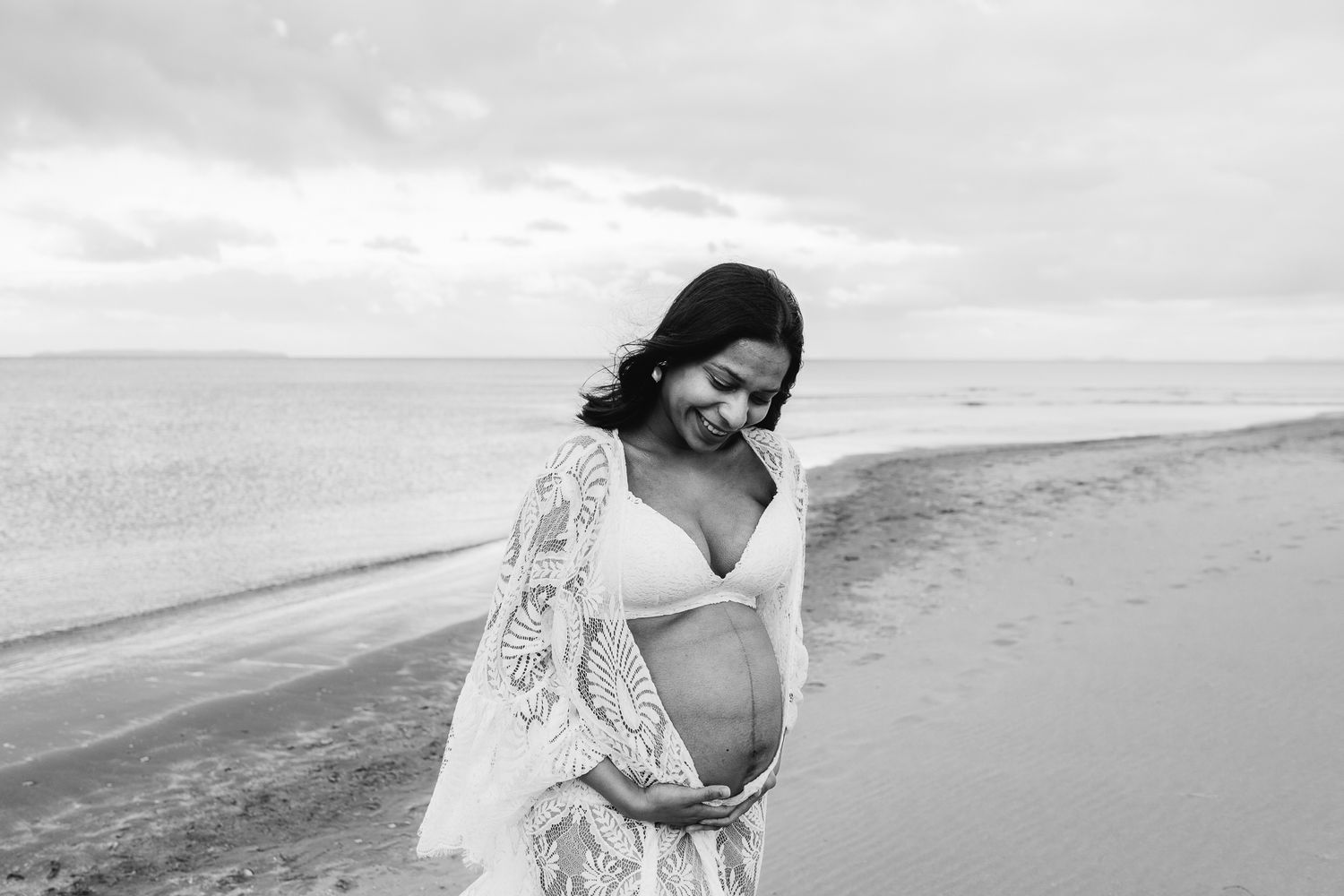 woman is wearing a white dress and is holding her baby belly as the sun sets behind her on the beach