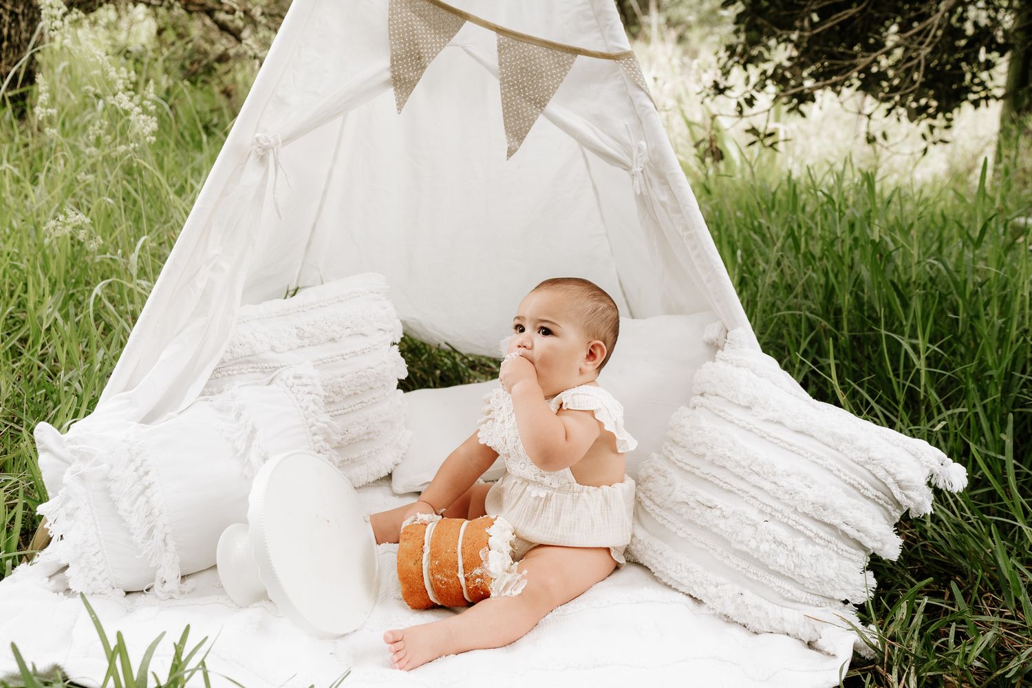 Outdoor cake smash session with a baby seated in front of a teepee, surrounded by long grass. A whimsical, natural setting with playful, mes