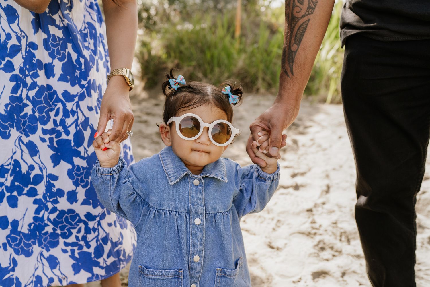 young girl with sunglasses on looks at the camera whilst mum and dad hold her hand
