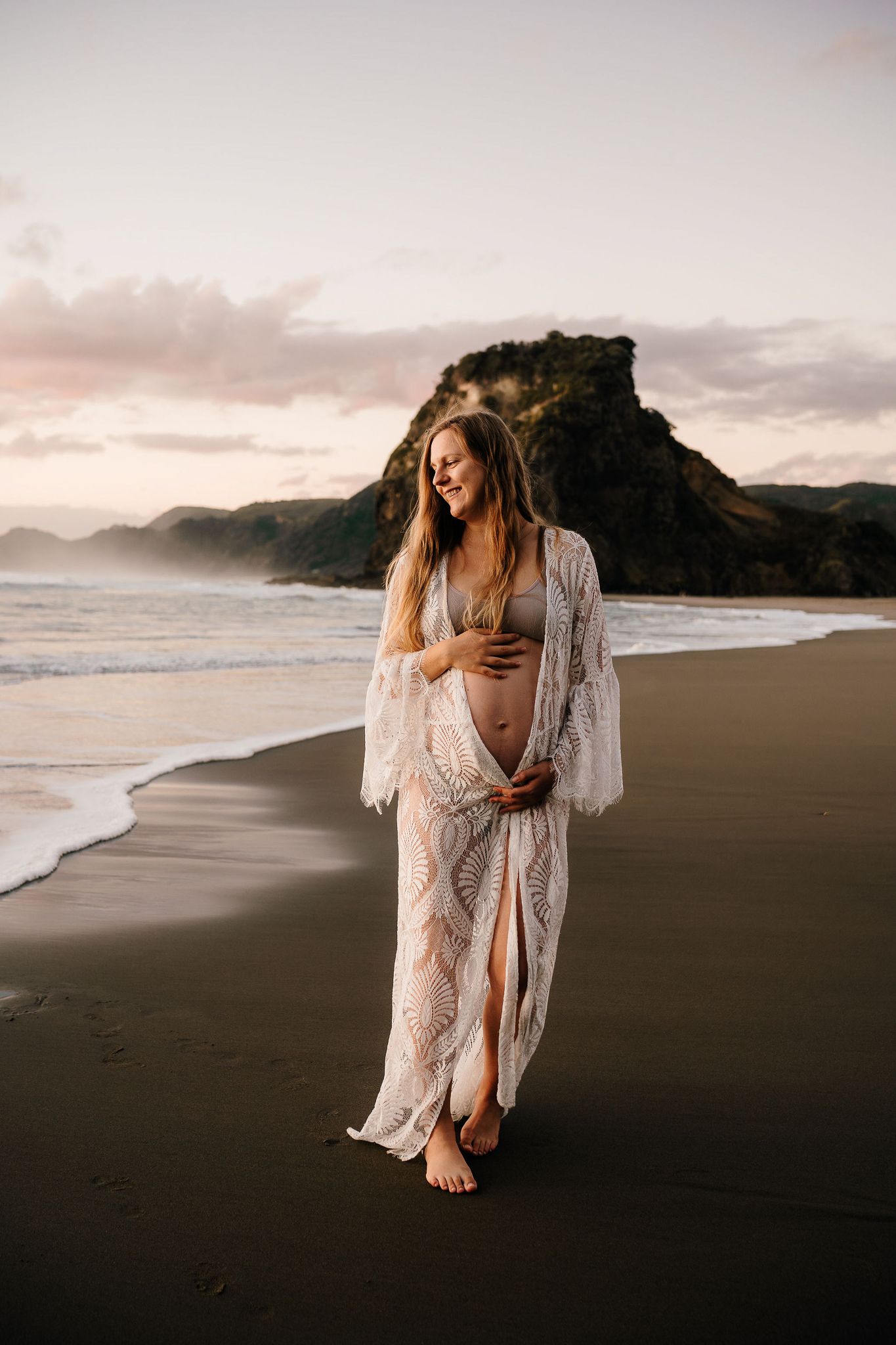 woman is wearing a white dress and is holding her baby belly as the sun sets behind her on the beach