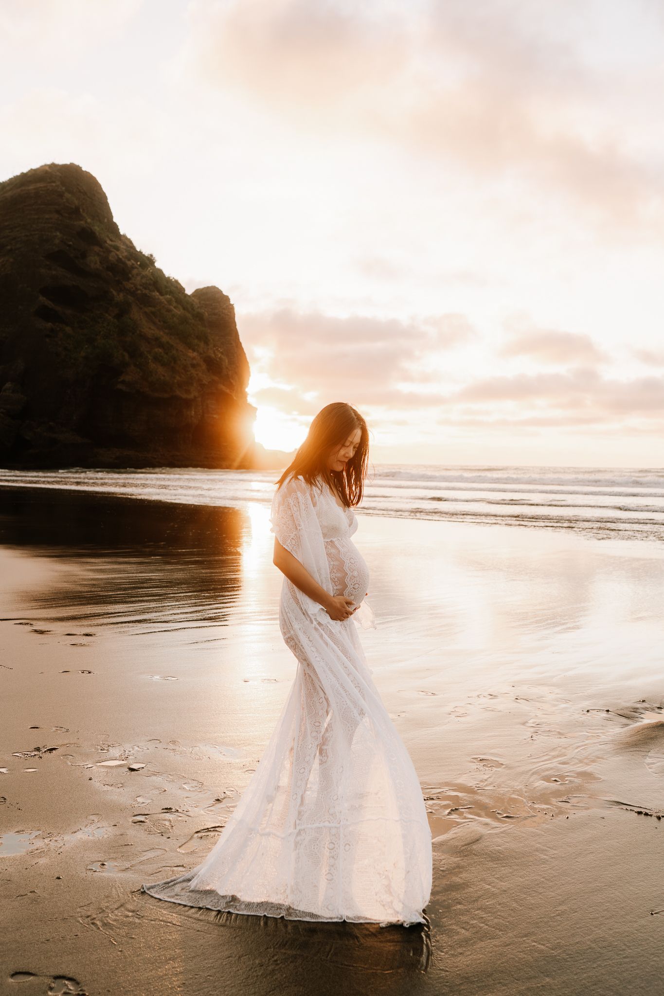 woman is wearing a white dress and is holding her baby belly as the sun sets behind her on the beach