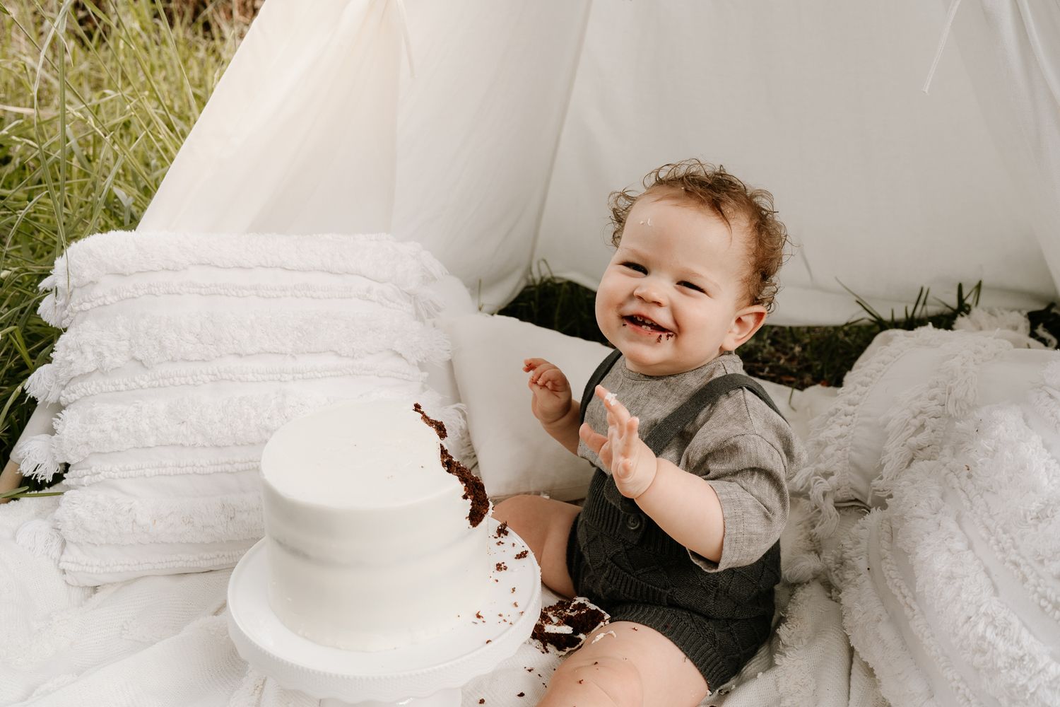 Outdoor cake smash session with a baby seated in front of a teepee, surrounded by long grass. A whimsical, natural setting with playful, mes