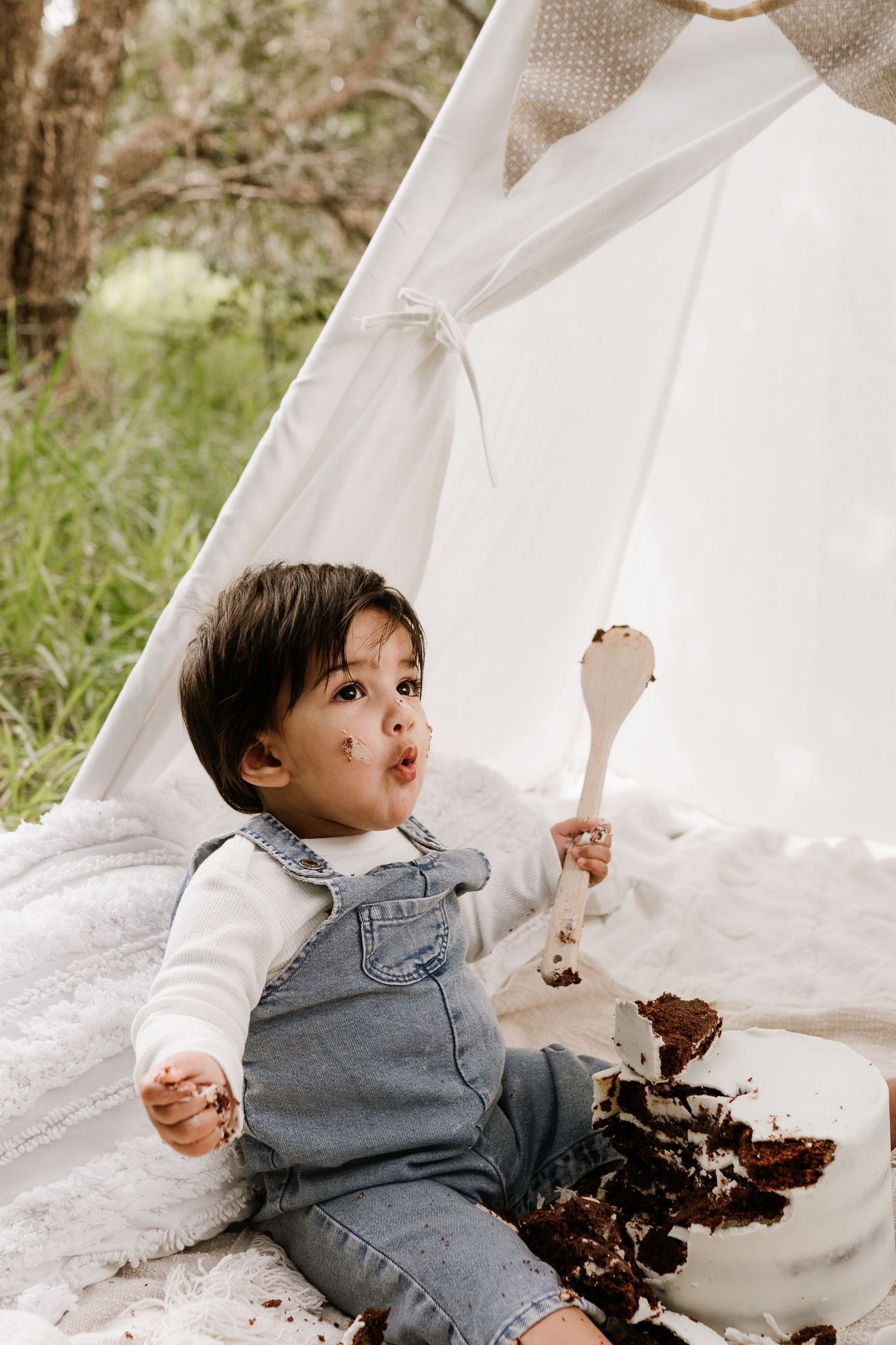 Outdoor cake smash session with a baby seated in front of a teepee, surrounded by long grass. A whimsical, natural setting with playful, mes