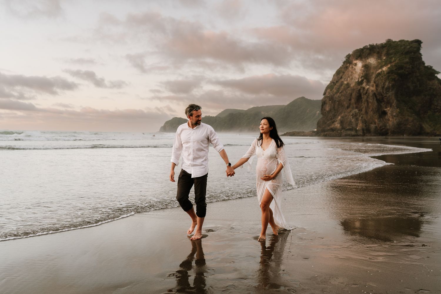 man and pregnant woman walk hand in hand along a dramatic New Zealand beach